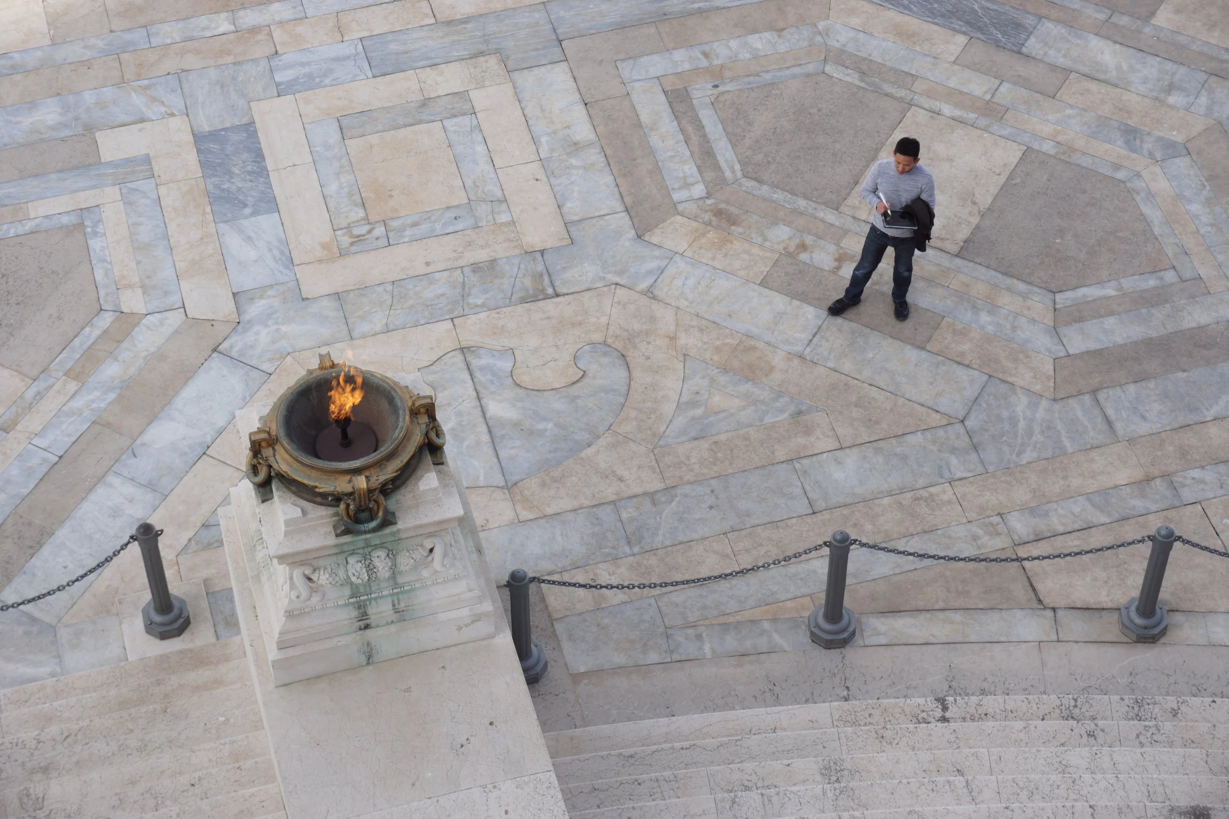 A person standing on a geometric paved area near a fire in an ornate stone fire bowl, viewed from above.