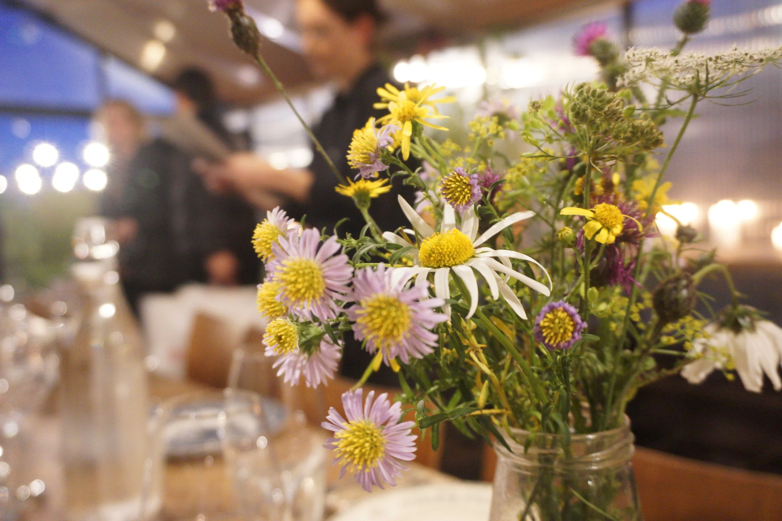 A close-up of a vase with wildflowers including daisies and other small purple and yellow flowers, on a table in a restaurant or cafe setting with blurred figures and warm lighting in the background.