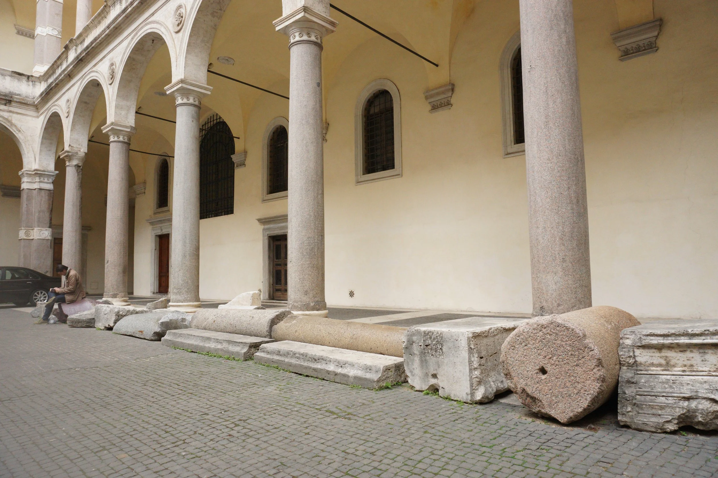 Ancient stone columns and ruins are displayed outside a historic building with arched windows and a yellow facade.