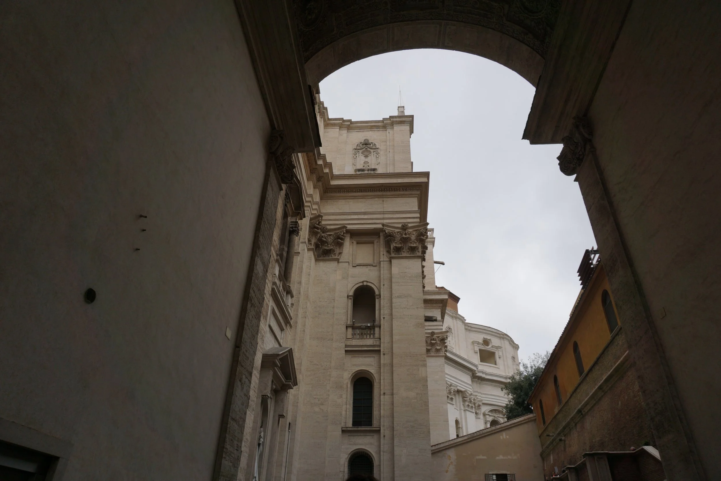View of a tall historic church or cathedral through an archway, with intricate architectural details and a cloudy sky above.