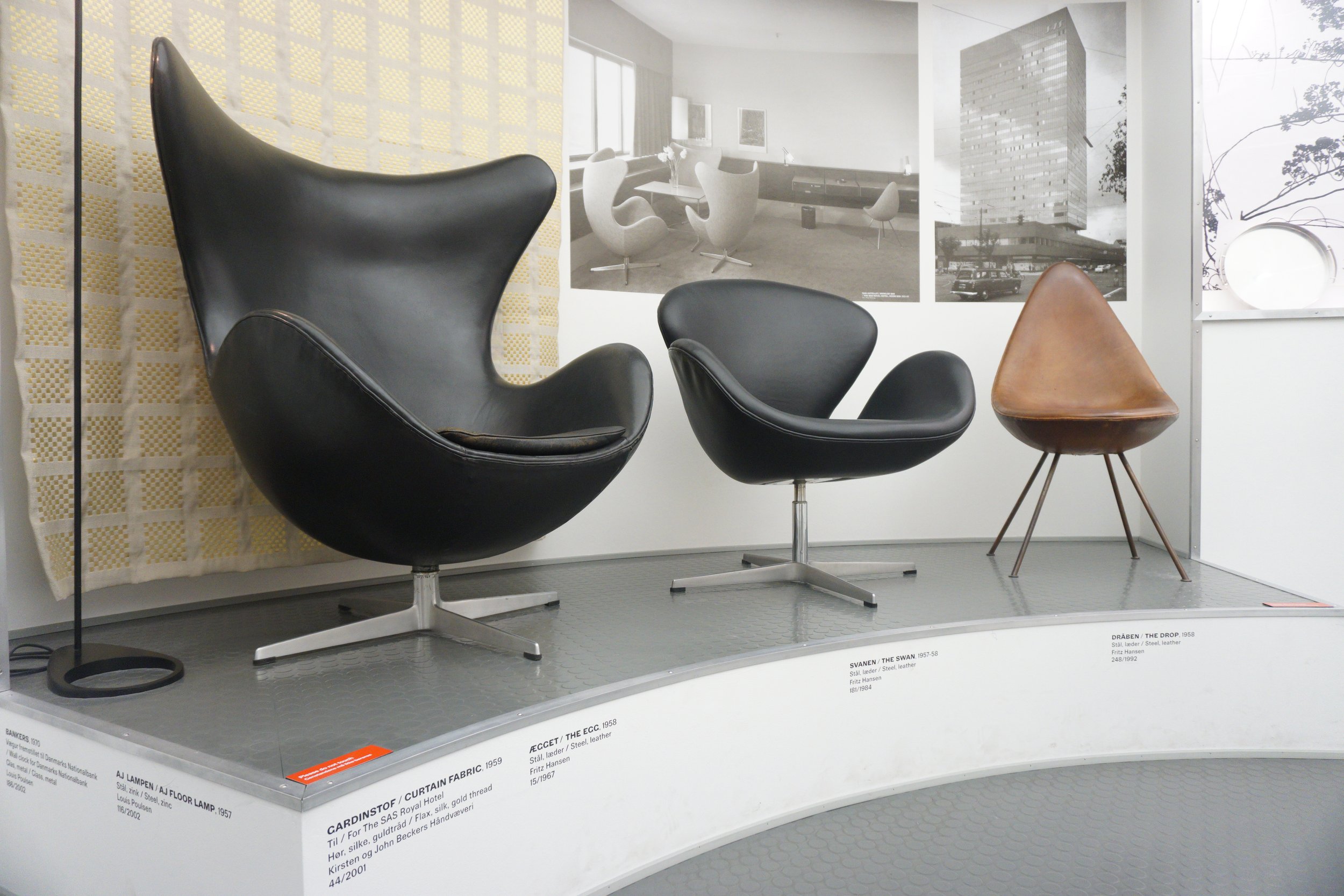 Three mid-century modern chairs displayed on a rounded platform with textual information, with black-and-white photos of buildings and furniture designs on the wall behind them.