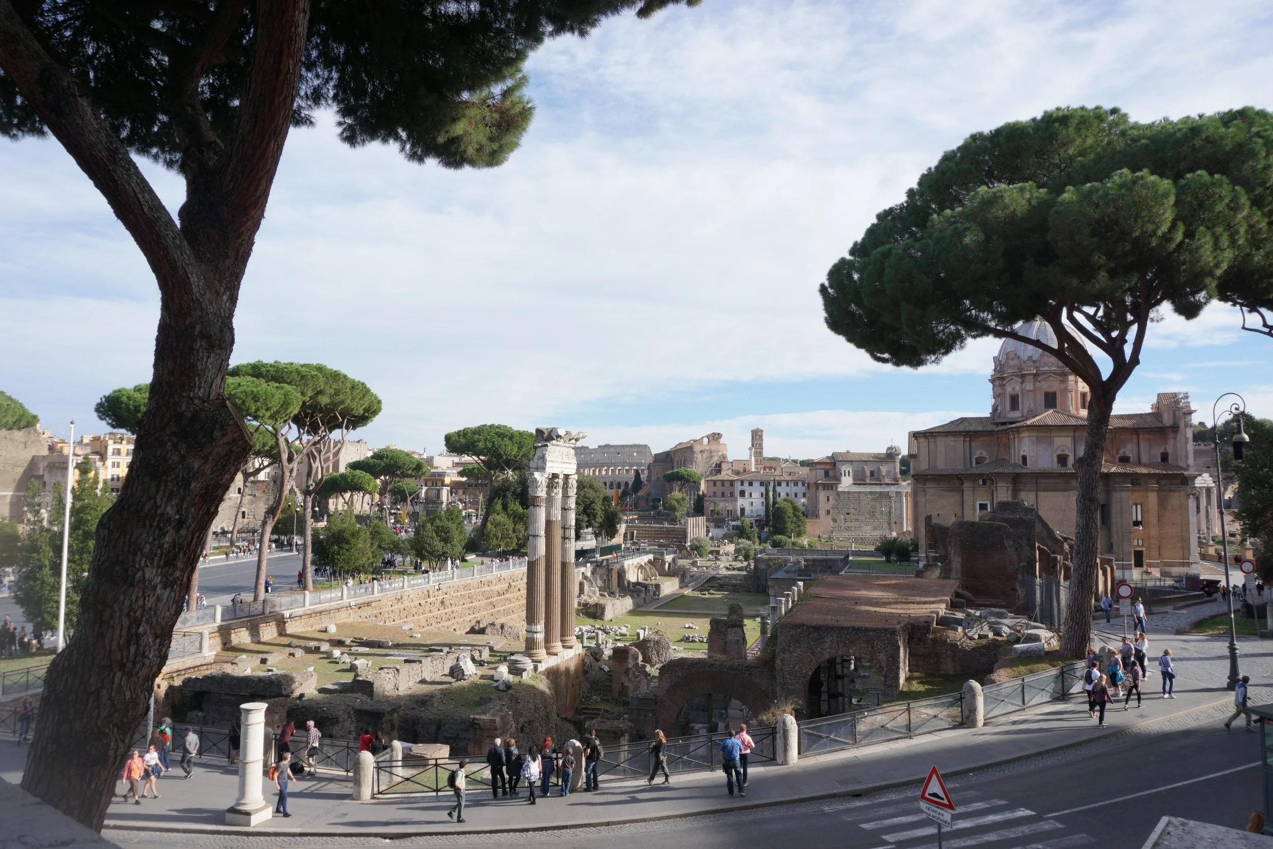 Ancient ruins and a church in Rome, Italy, with trees and people walking on the street.
