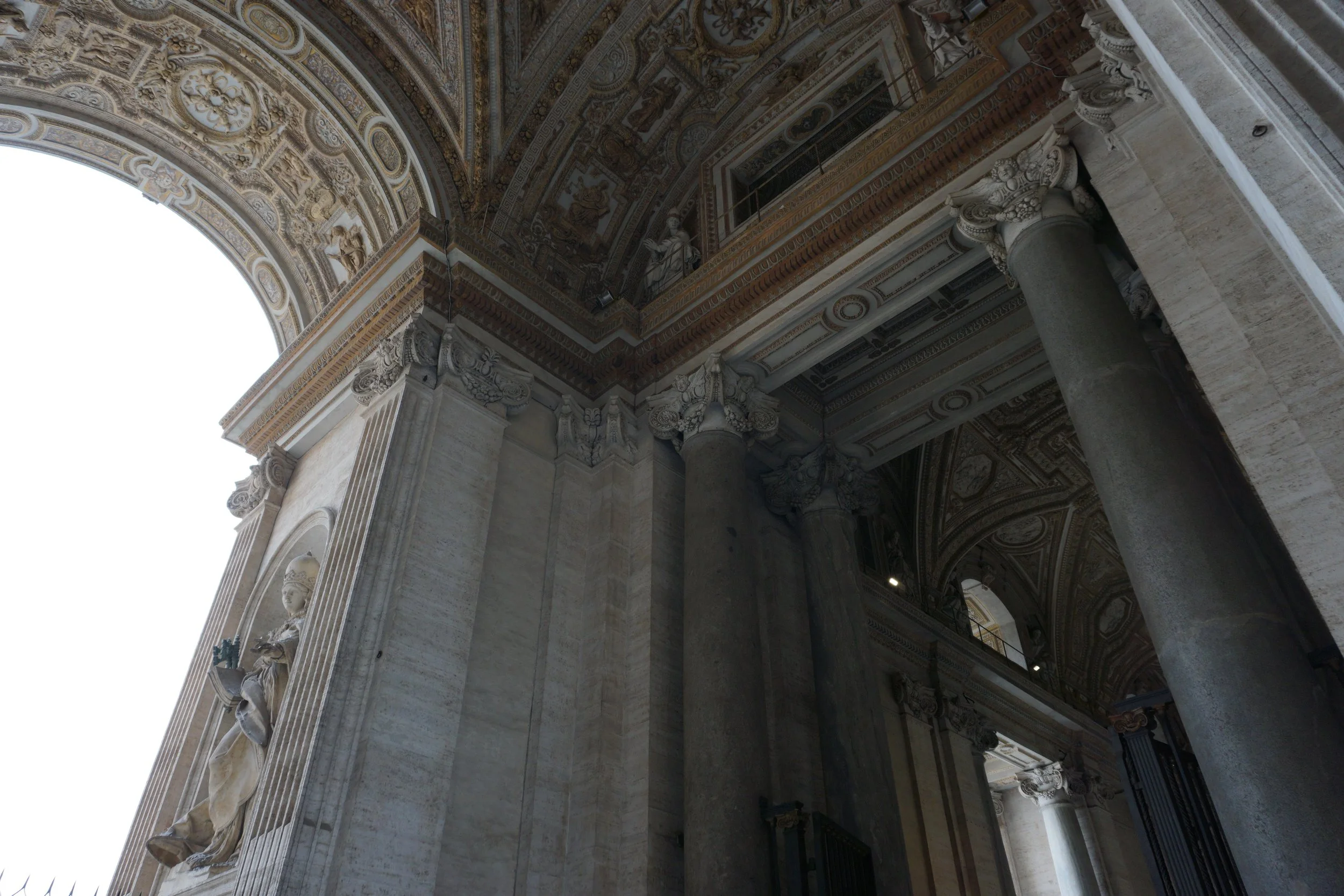 Interior view of an ornate classical building with large stone columns, detailed carvings, and statues. The ceiling features intricate patterns and artwork with gold accents.