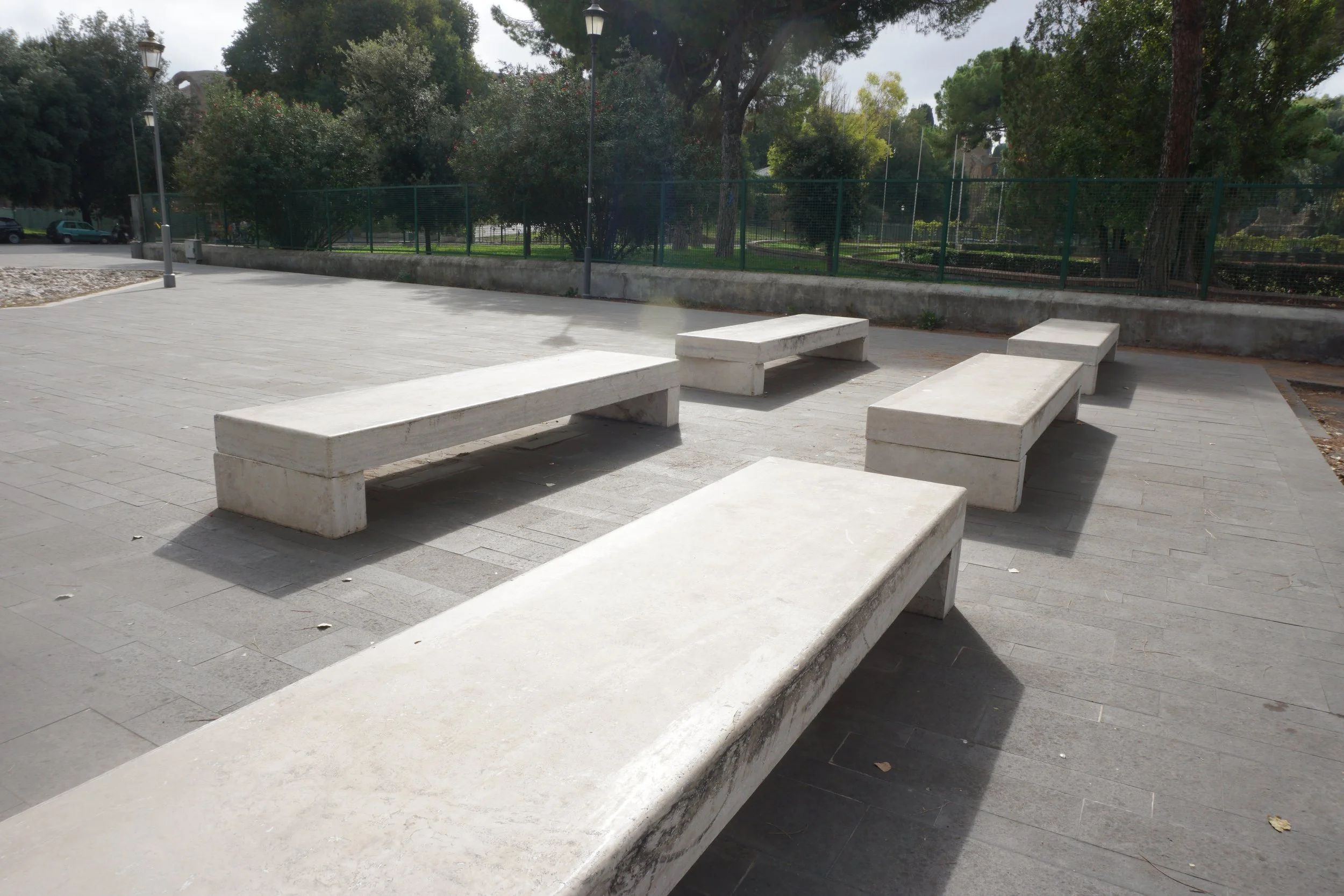 Four white concrete benches are arranged on a paved outdoor area, with a green park and trees in the background.