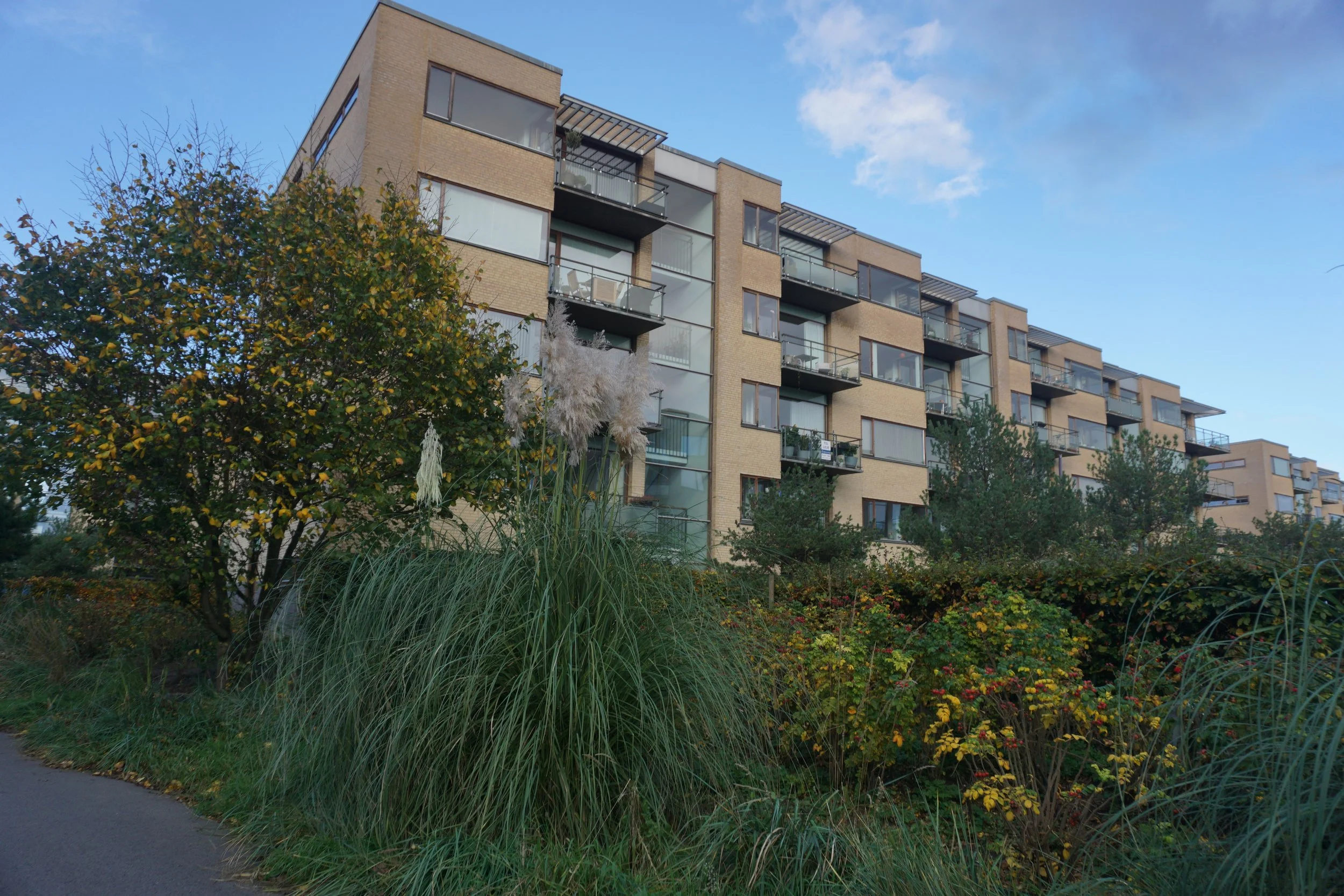 Modern multi-story apartment building with glass balconies, surrounded by green trees and shrubs, under a partly cloudy sky.