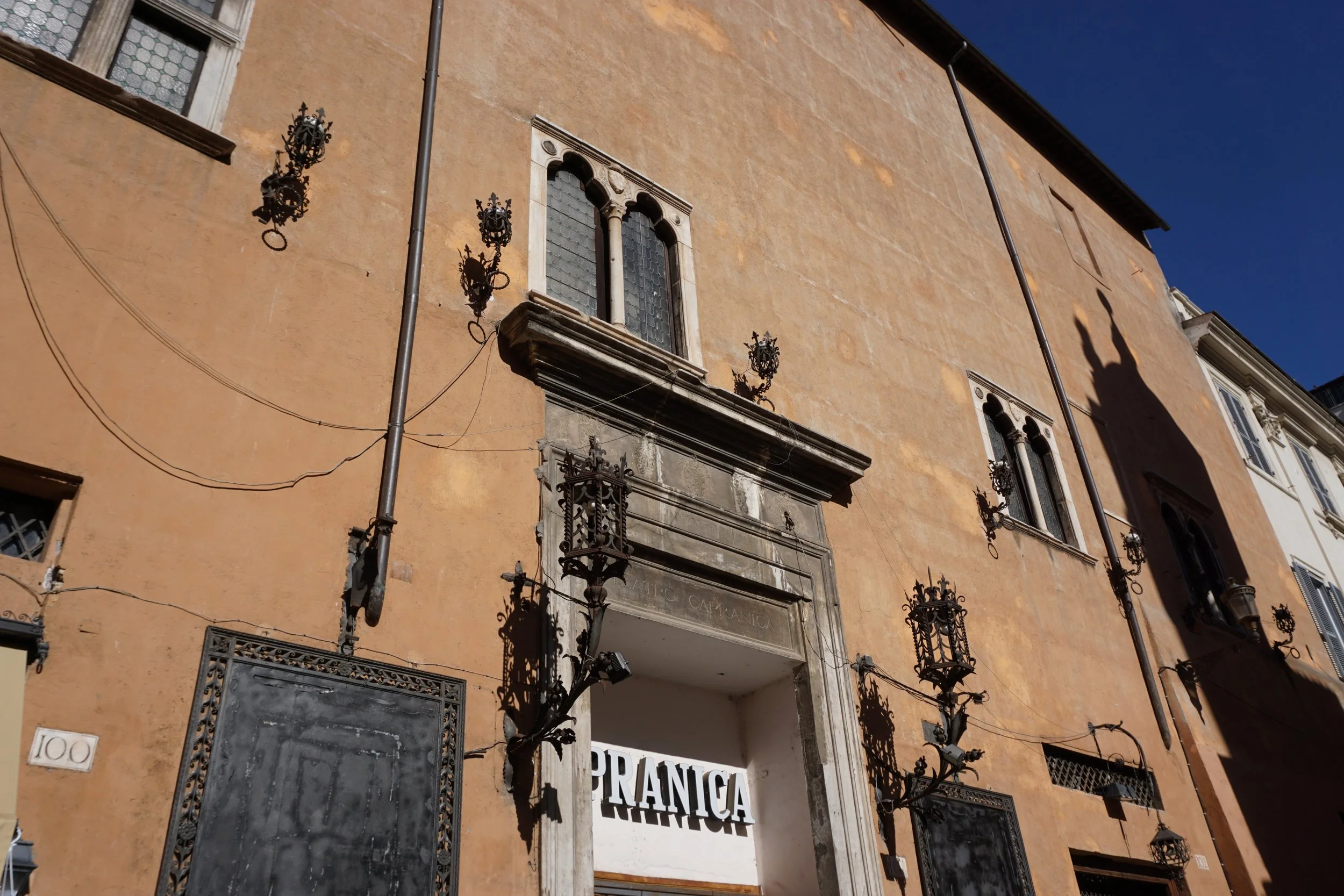 View of an old building with a peach-colored facade, decorative windows, black wrought iron lanterns, and an archway entrance with a sign that reads 'TRARNICA'.