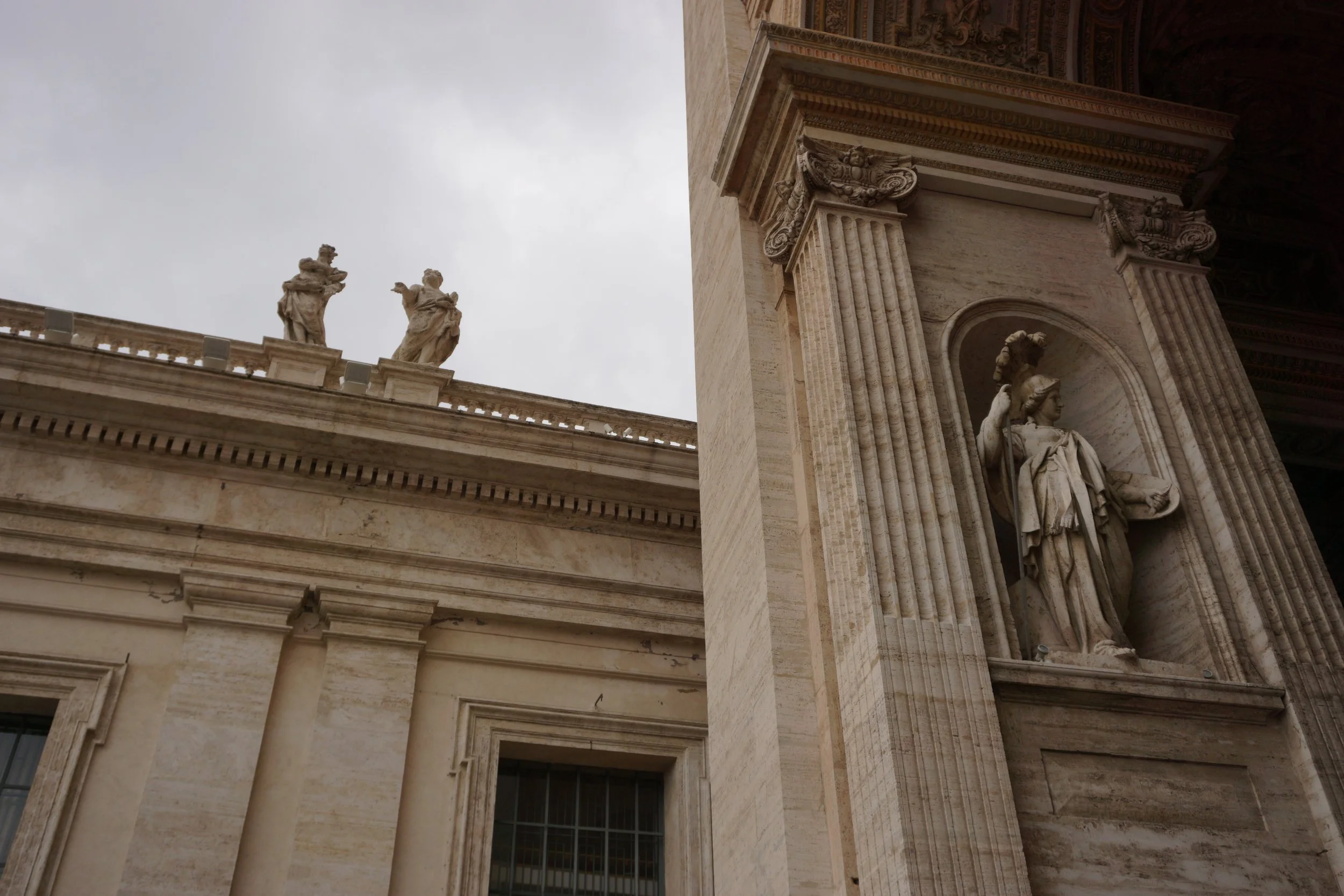 Close-up of classical architecture with ornate statues on a building, showing detailed stone sculptures and columns against an overcast sky.