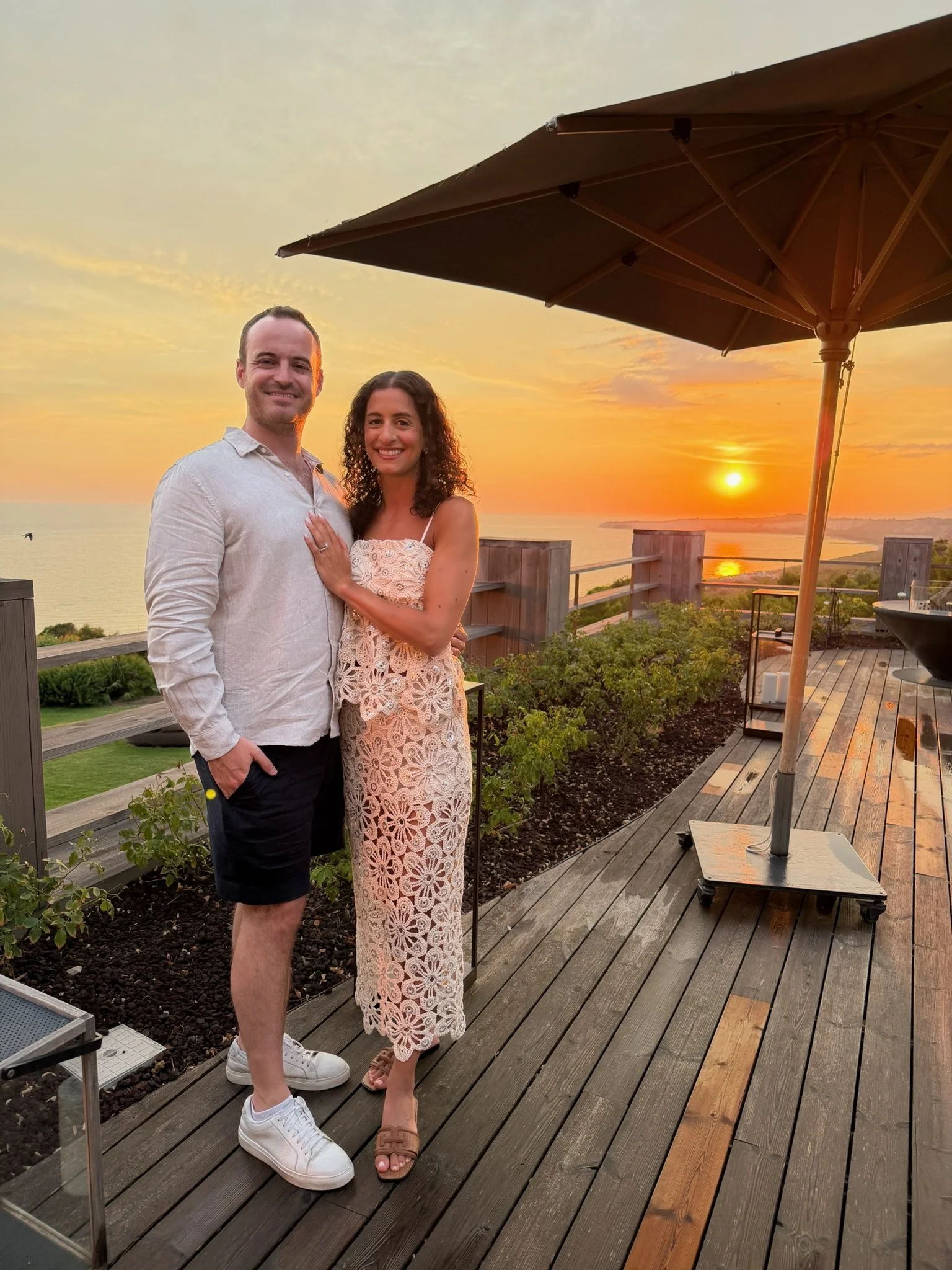 Alexia and Dan embracing on a terrace overlooking the Sicilian coastline at sunset.