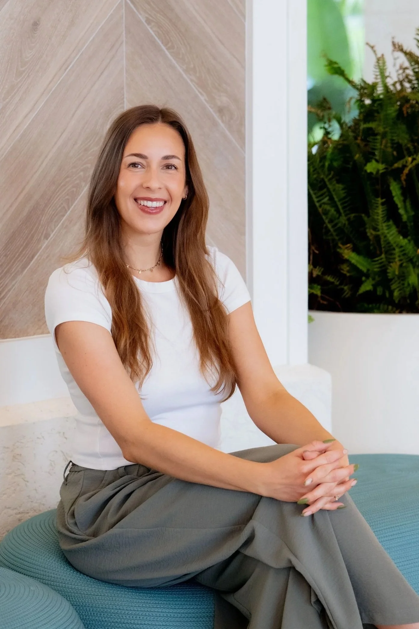 a woman in her 30s with long brown hair wearing a white t-shirt and green pants poses for a professional photo, smiling