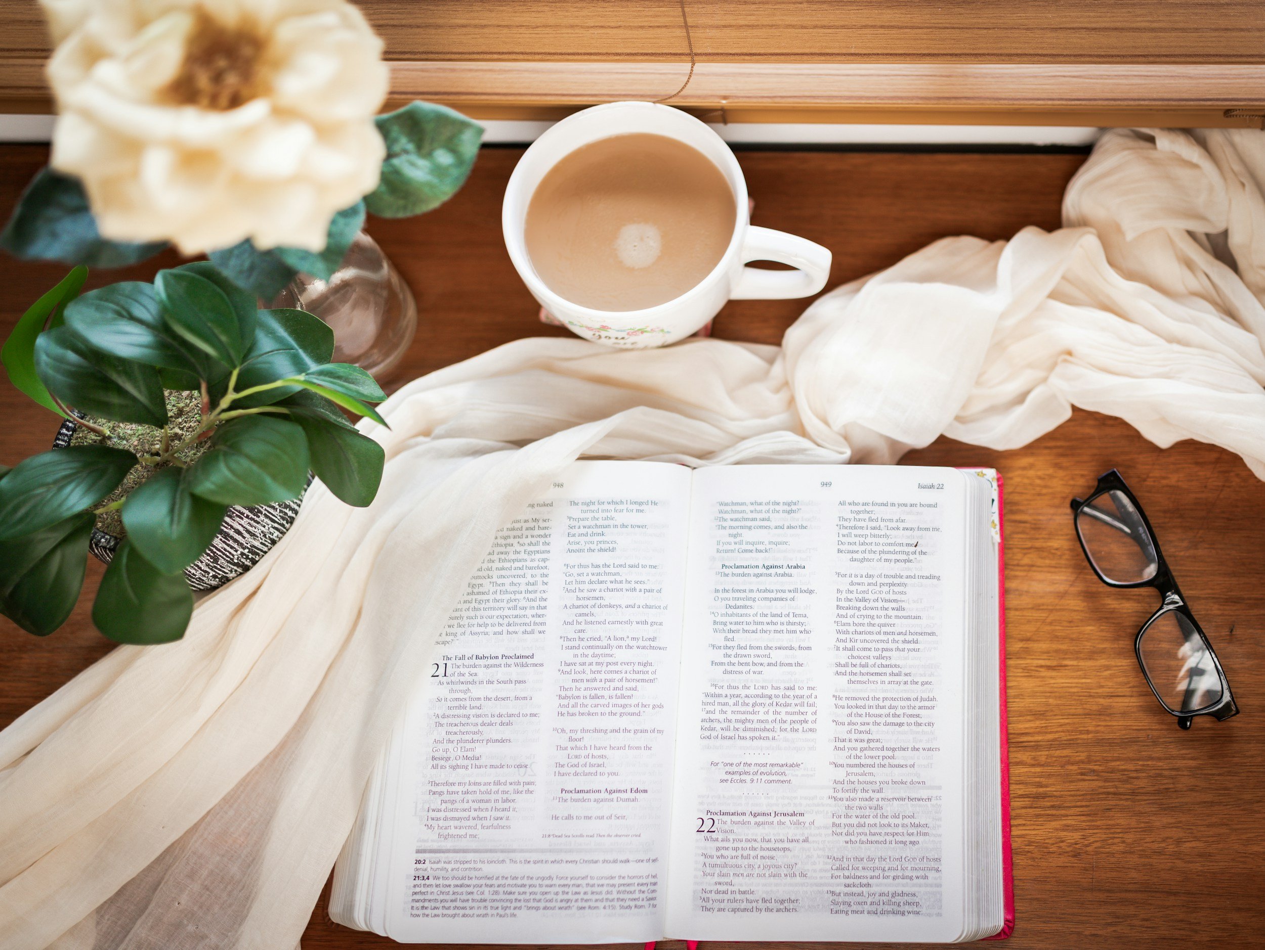 Open Bible with text pages, a white mug of coffee, a white cloth, a potted green plant, a flower in a vase, and a pair of glasses on a wooden table.