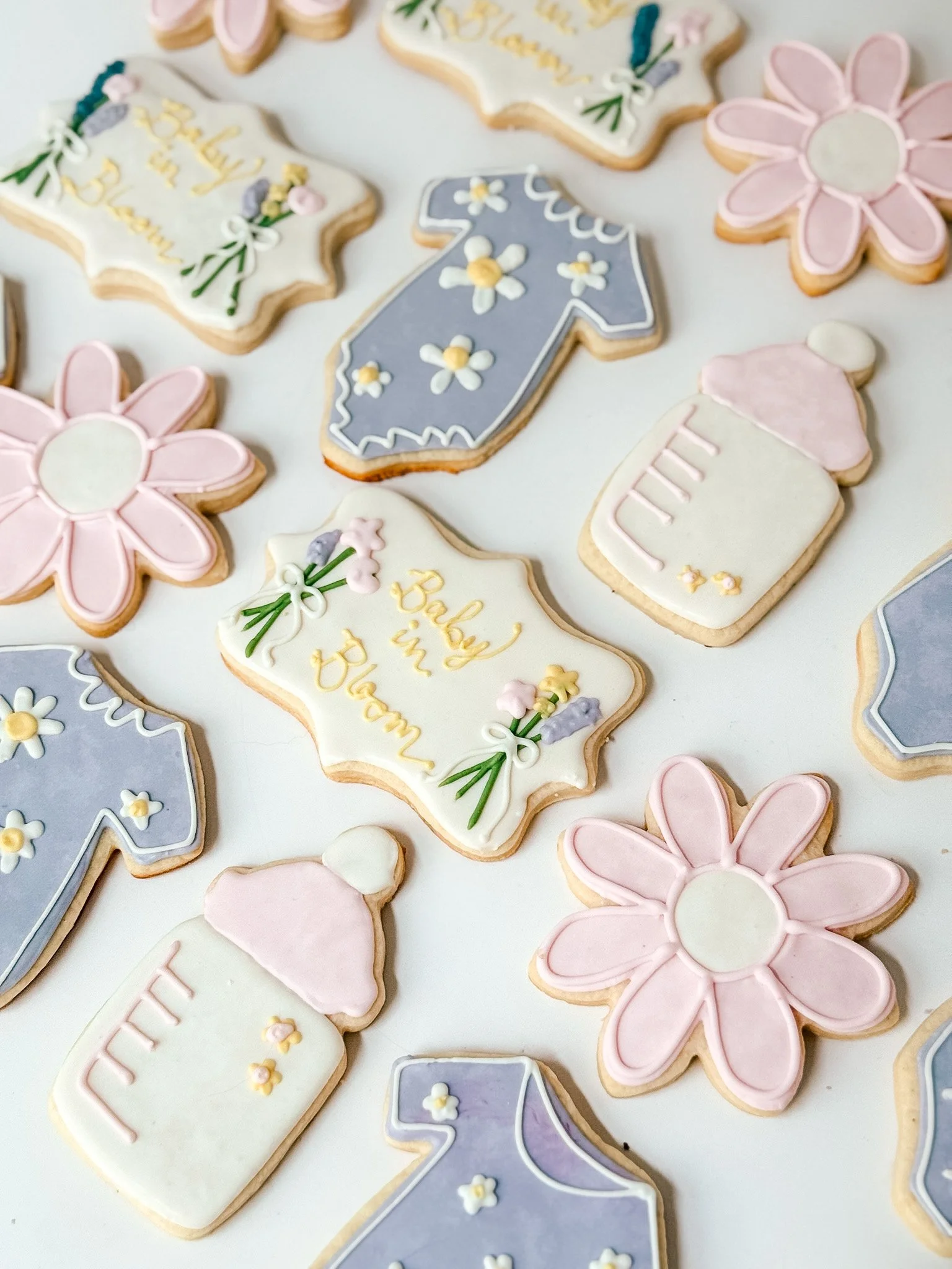 Assorted decorated cookies in floral, baby bottle, gift, and sock topics with pastel colors, on a white background.
