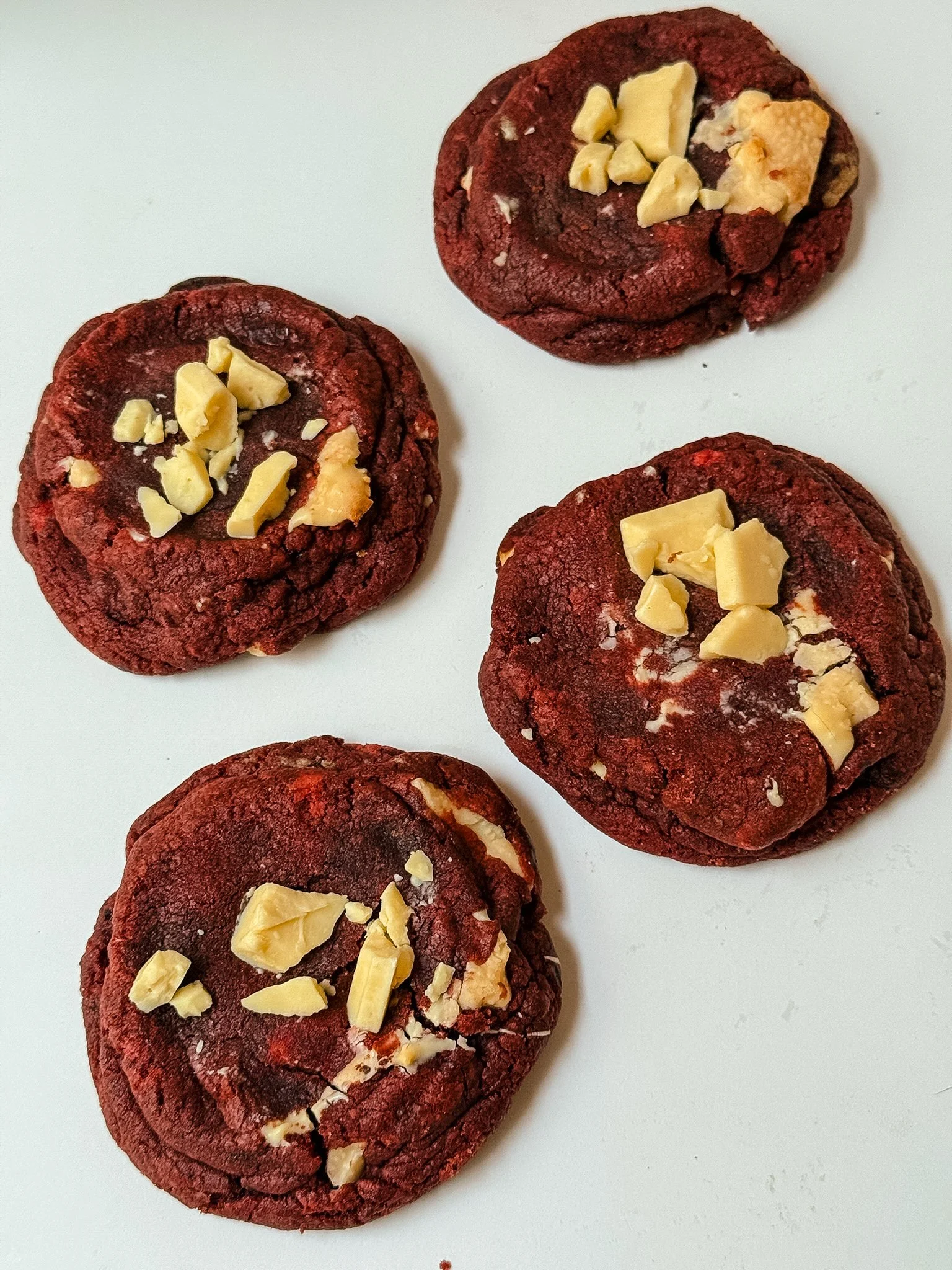 Close-up of five red velvet cookies topped with chopped white chocolate pieces on a white surface.