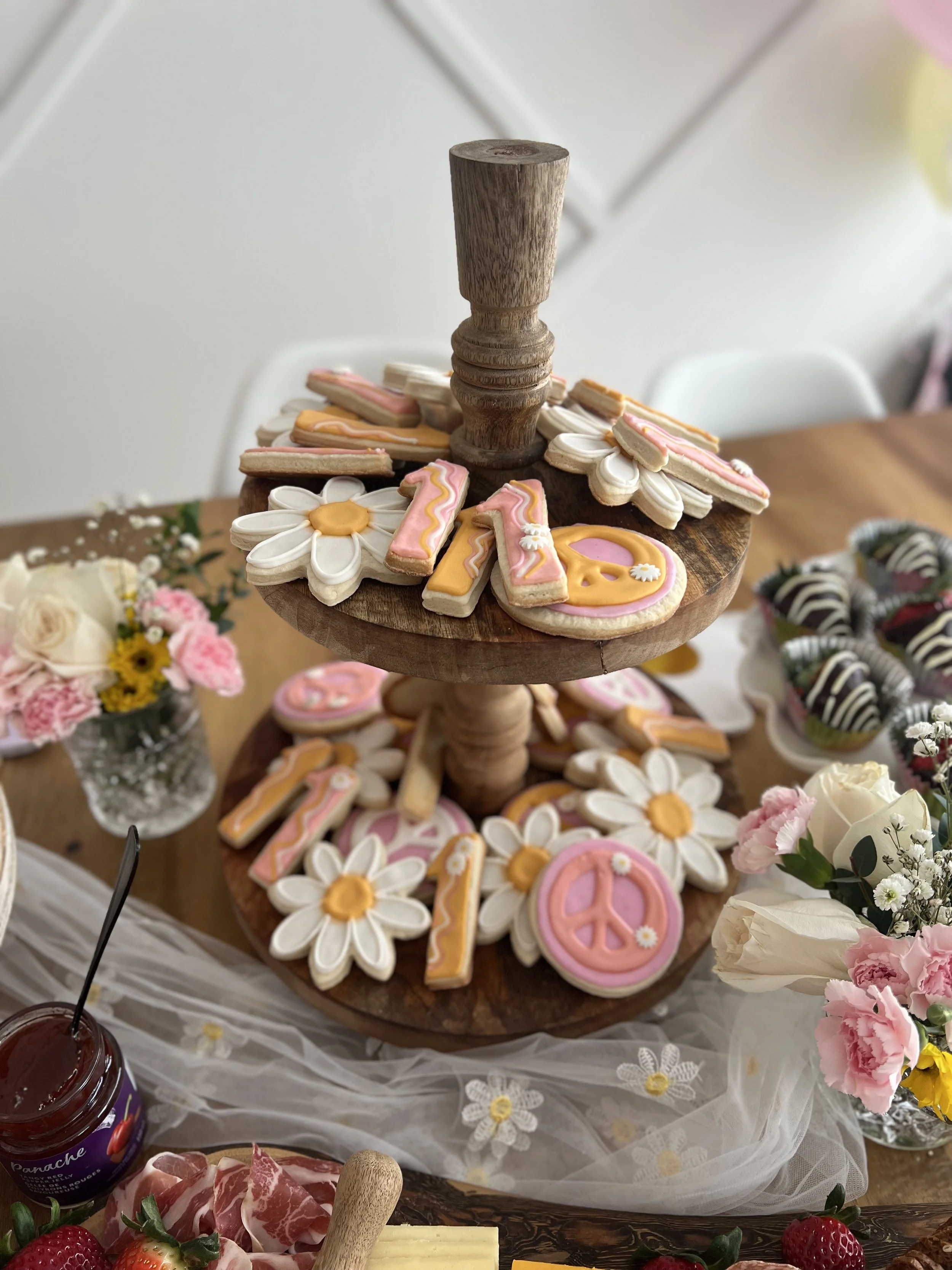 A two-tier wooden serving stand with decorated sugar cookies in various shapes, including flowers, peace signs, and letters, on a table. Surrounding the stand are flowers, strawberries, and other breakfast items.