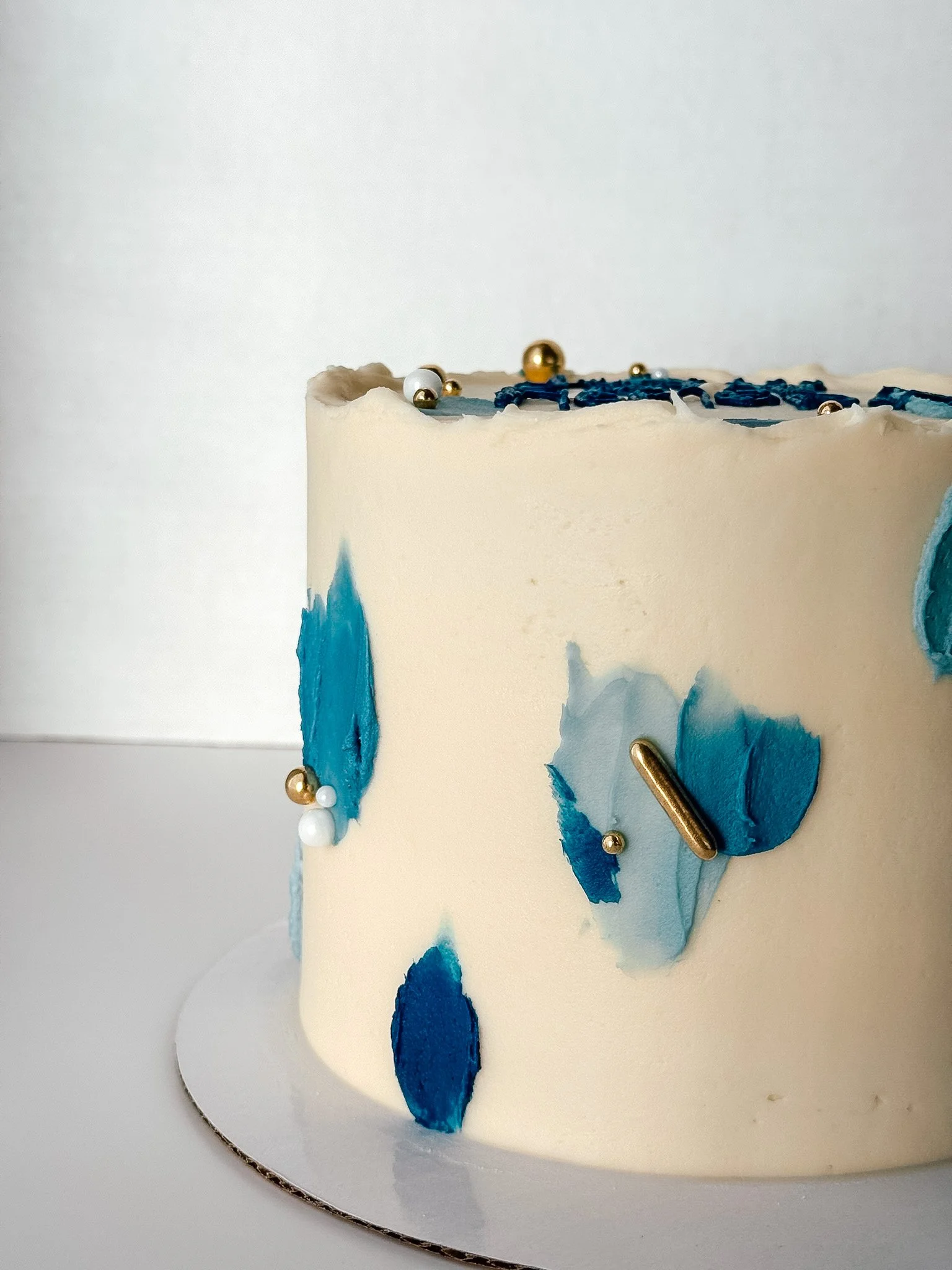 A white cake decorated with blue paint, white and gold spherical beads, and a gold metallic object, placed on a white plate.