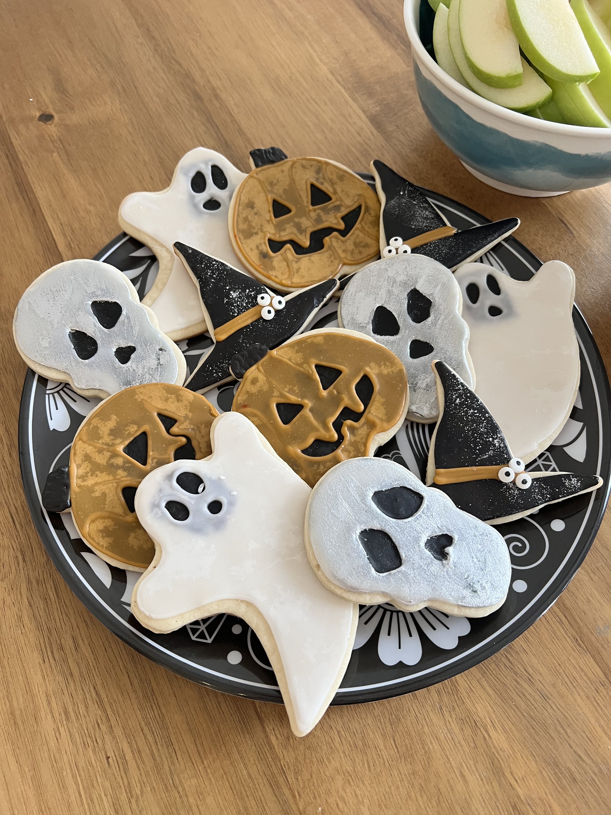 Halloween-themed cookies shaped like ghosts, pumpkins, and black witch hats with small googly eyes on a black and white patterned plate, next to a bowl of sliced green apples on a wooden table.