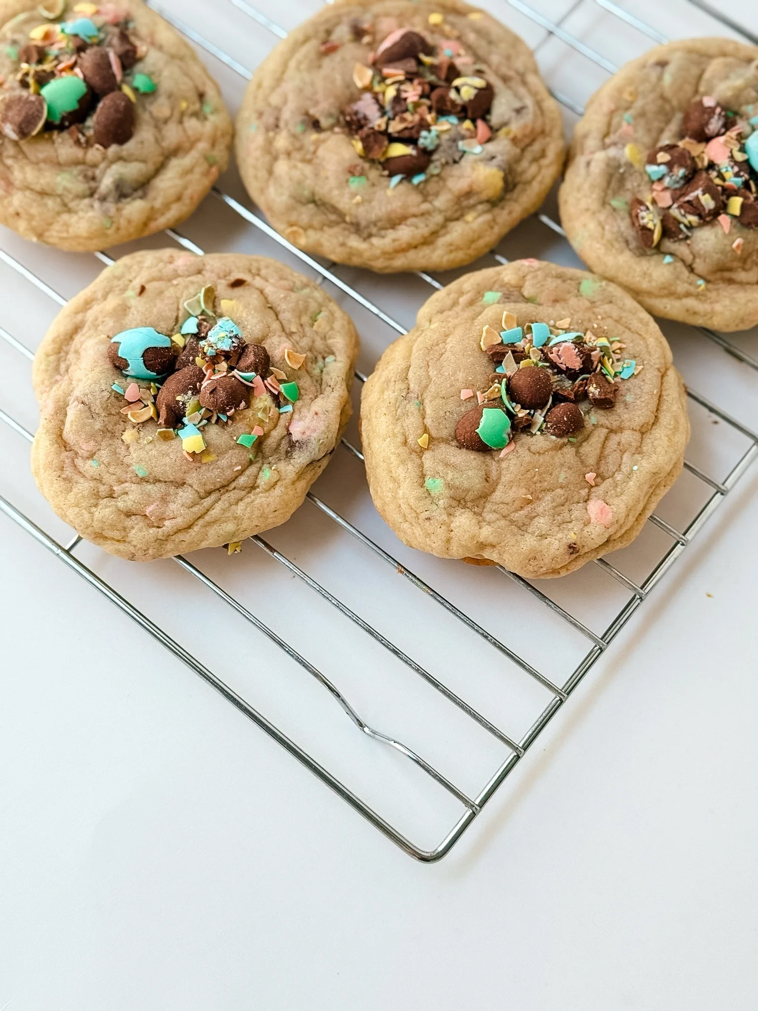 Five cookies with chocolate chunks and colorful sprinkles cooling on a metal wire rack.