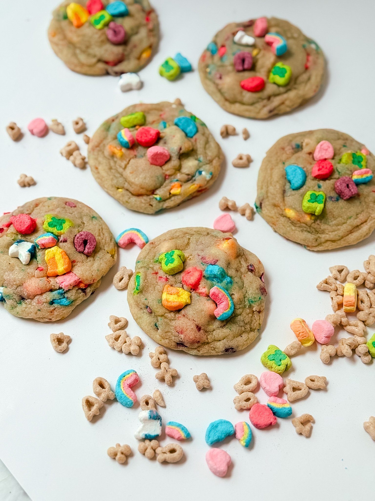 Group of colorful cookies decorated with rainbow and heart-shaped sprinkles, scattered on a white surface along with alphabet-shaped cereal and small heart-shaped candies.