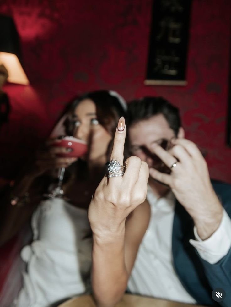 A woman with long brown hair holding a glass of red wine and showing her middle finger, which has a large diamond ring, in front of a man with dark hair, wearing a suit, making a rock and roll hand gesture in a dimly lit red room.
