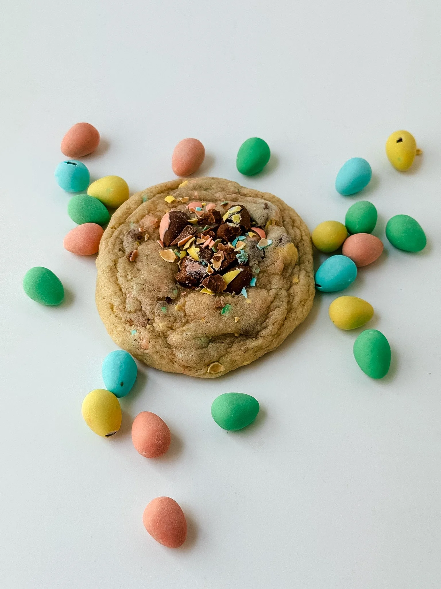 A cookie topped with chocolate chunks and colorful sprinkles, surrounded by pastel-colored candy eggs on a white background.