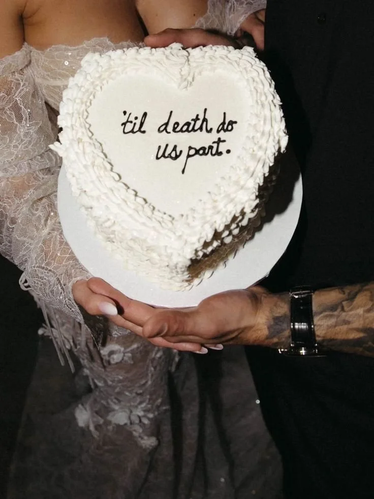 A couple holding a heart-shaped wedding cake with white frosting. The cake has the message '’til death do us part.' written on it in black icing.
