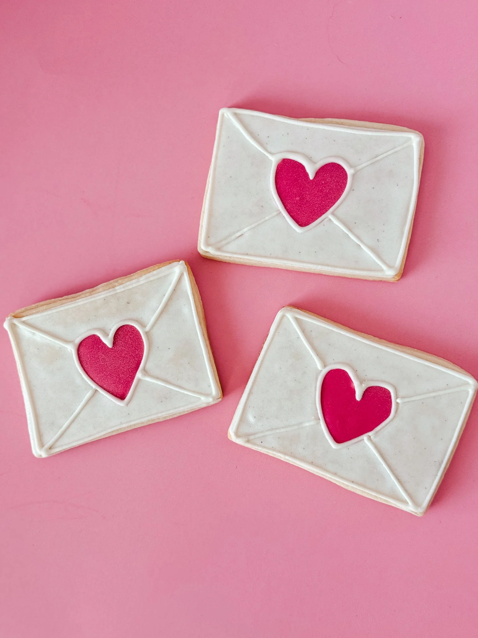 Decorated sugar cookies shaped like envelopes with pink hearts on top, placed on a pink surface.