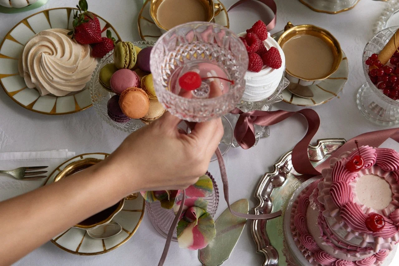 A table set for a celebration with a variety of desserts and drinks, including a pink frosted cake with cherries, macarons, strawberries, raspberries, red currants, and cups of tea or coffee, with a person holding a decorative glass with a cherry on top.
