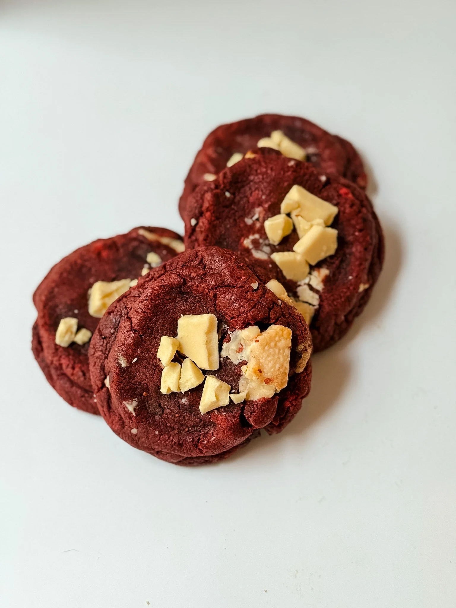 A close-up view of four red velvet cookies topped with white chocolate chunks on a white background.