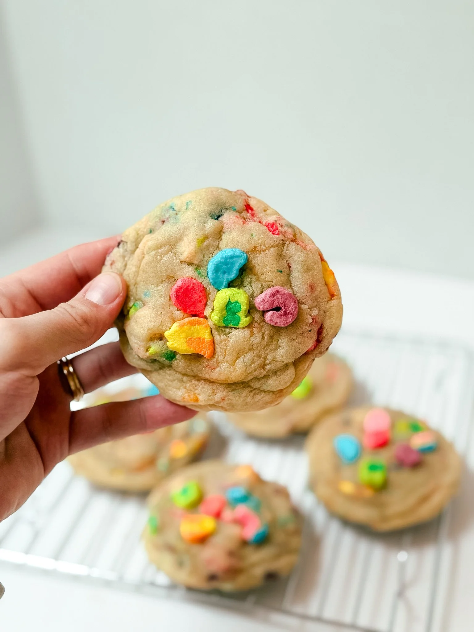 Hand holding a large sugar cookie decorated with colorful candy pieces, with more decorated cookies on a cooling rack in the background.