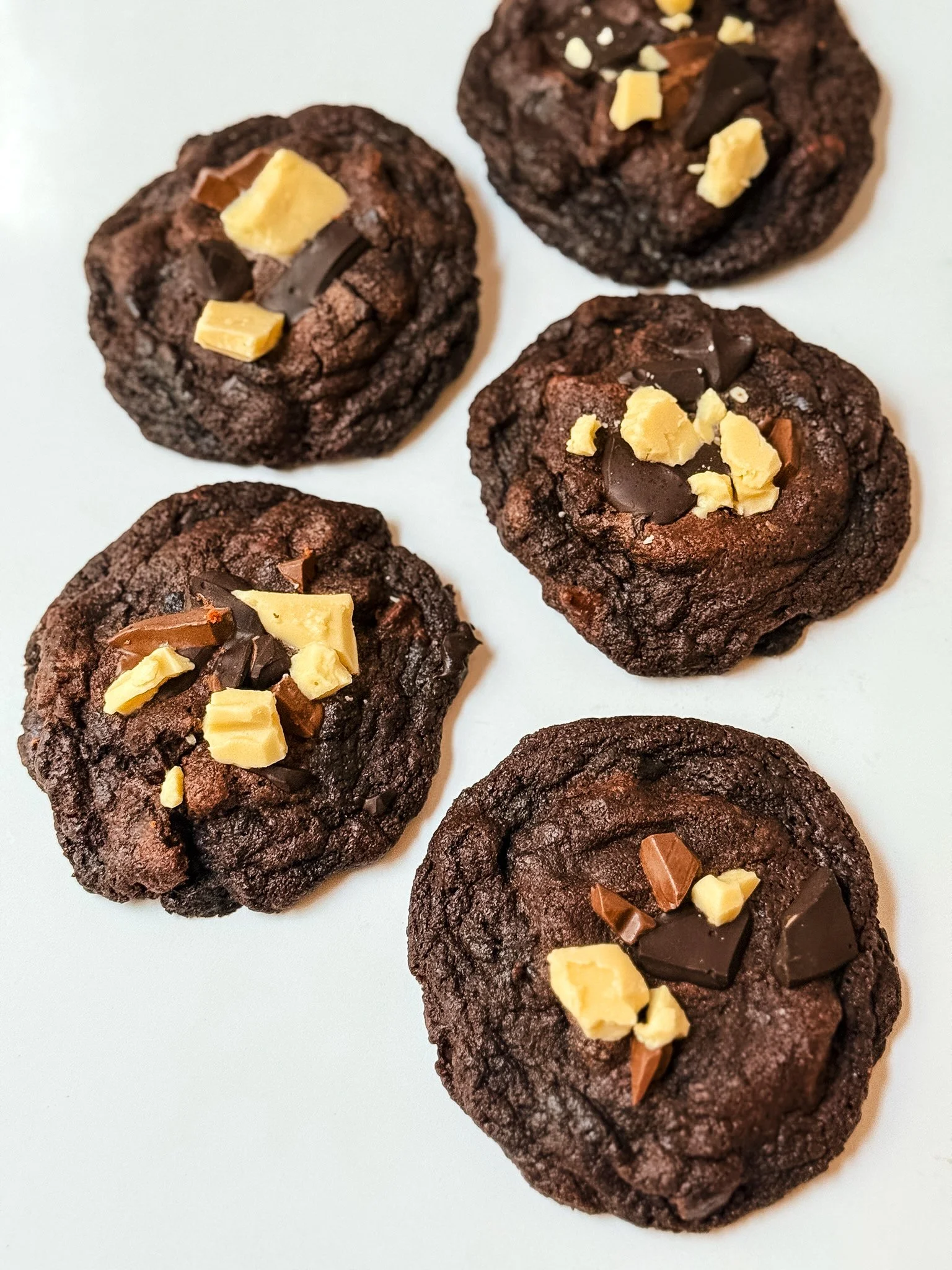 Close-up of five chocolate cookies topped with pieces of white chocolate and dark chocolate, arranged on a white surface.