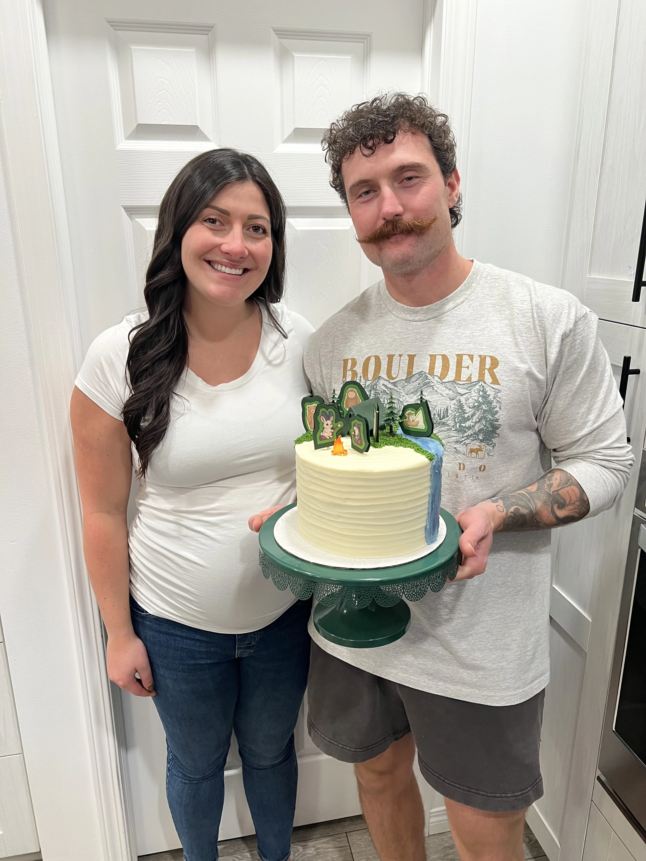 A woman and a man are standing together indoors, smiling at the camera. The woman has long dark hair, and the man has a curly hairstyle, beard, and a distinctive mustache. The man is holding a round cake with forest-themed toppers, including animals 