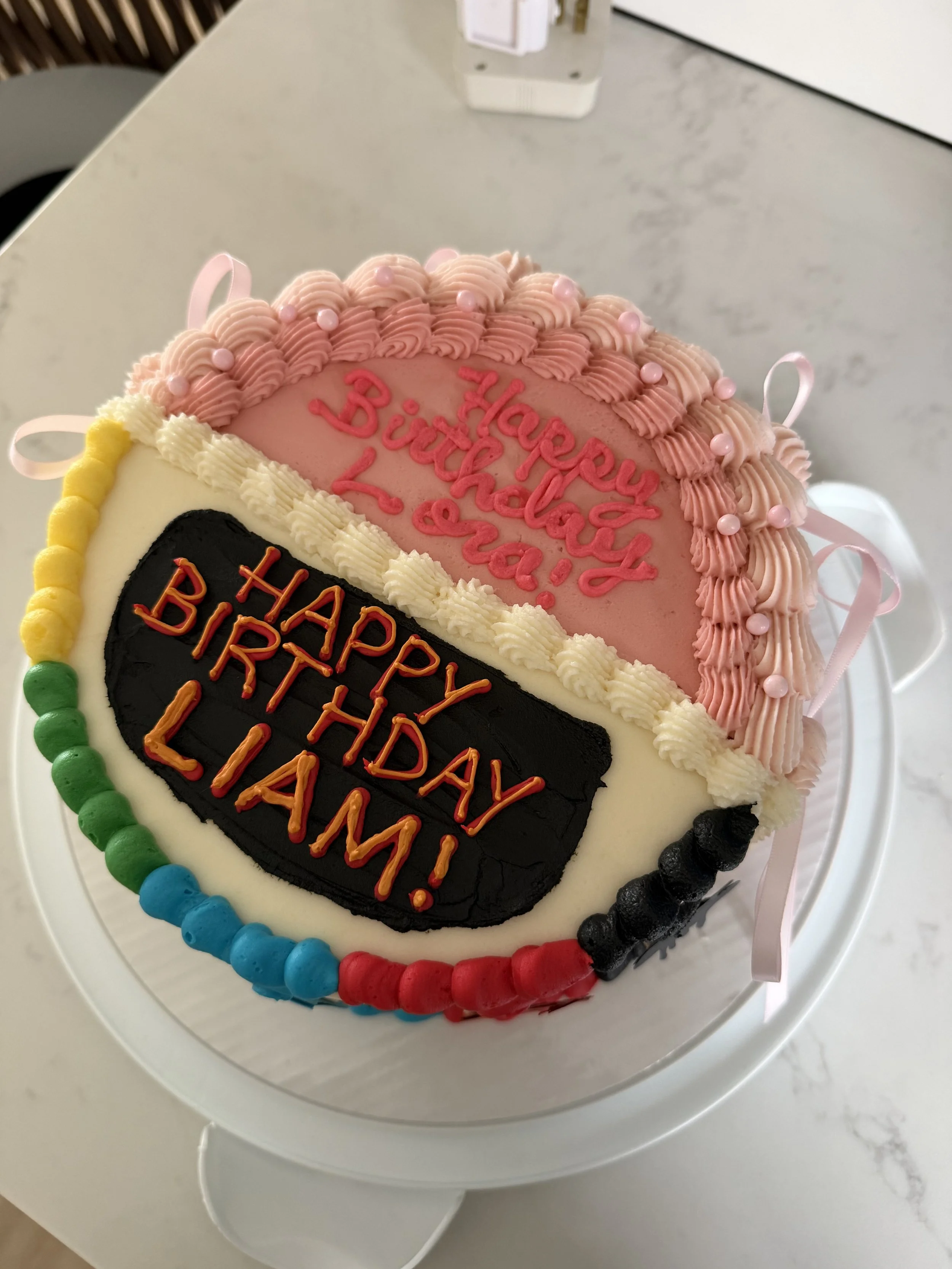 Colorful birthday cake with pink, white, green, and red decorations, topped with pink frosting, pink pearls, and a pink ribbon, with a message written in pink icing on top.