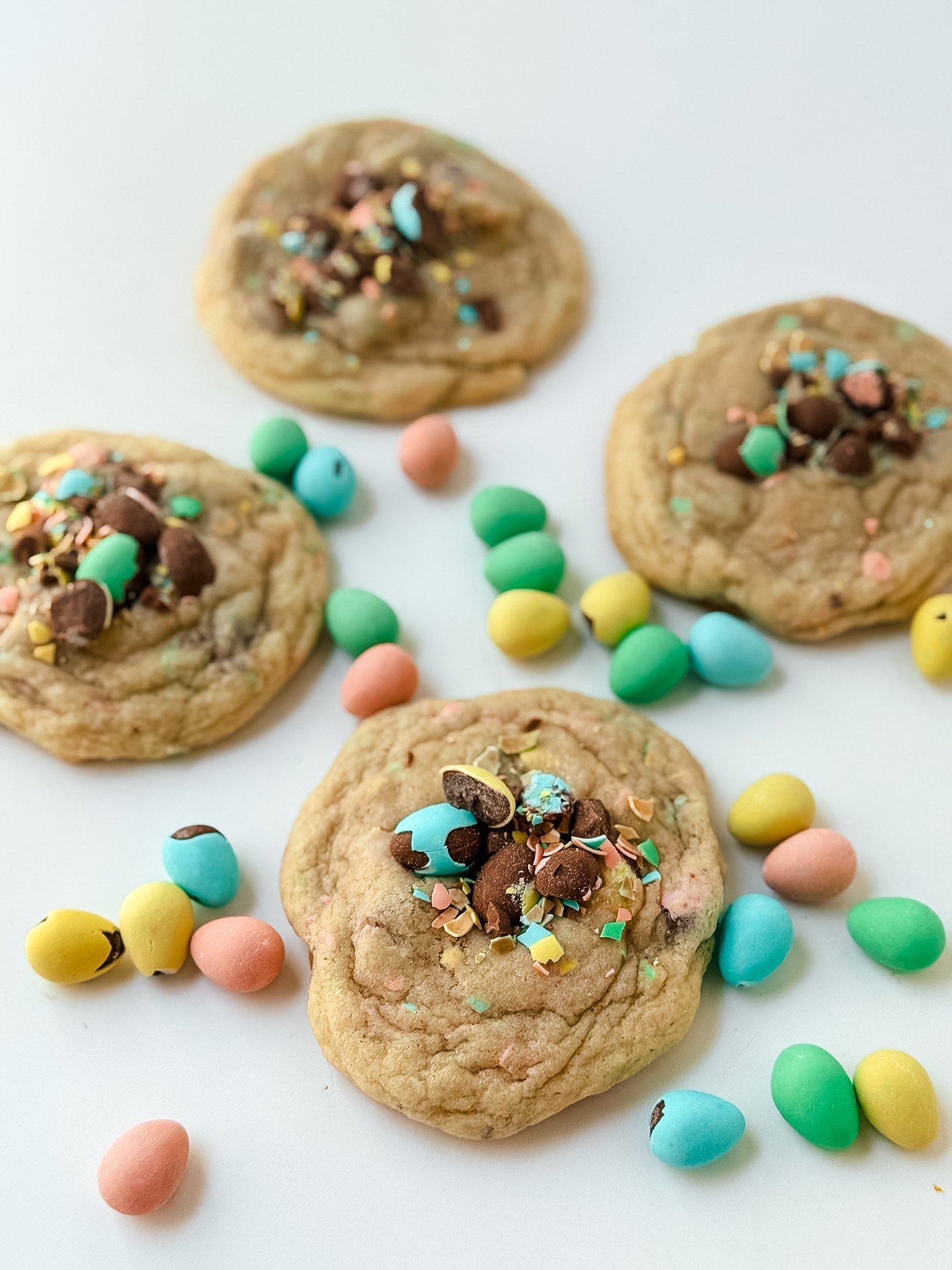 Four cookies decorated with colorful sprinkles, chocolate chips, and candies, surrounded by pastel-colored candies on a white background.