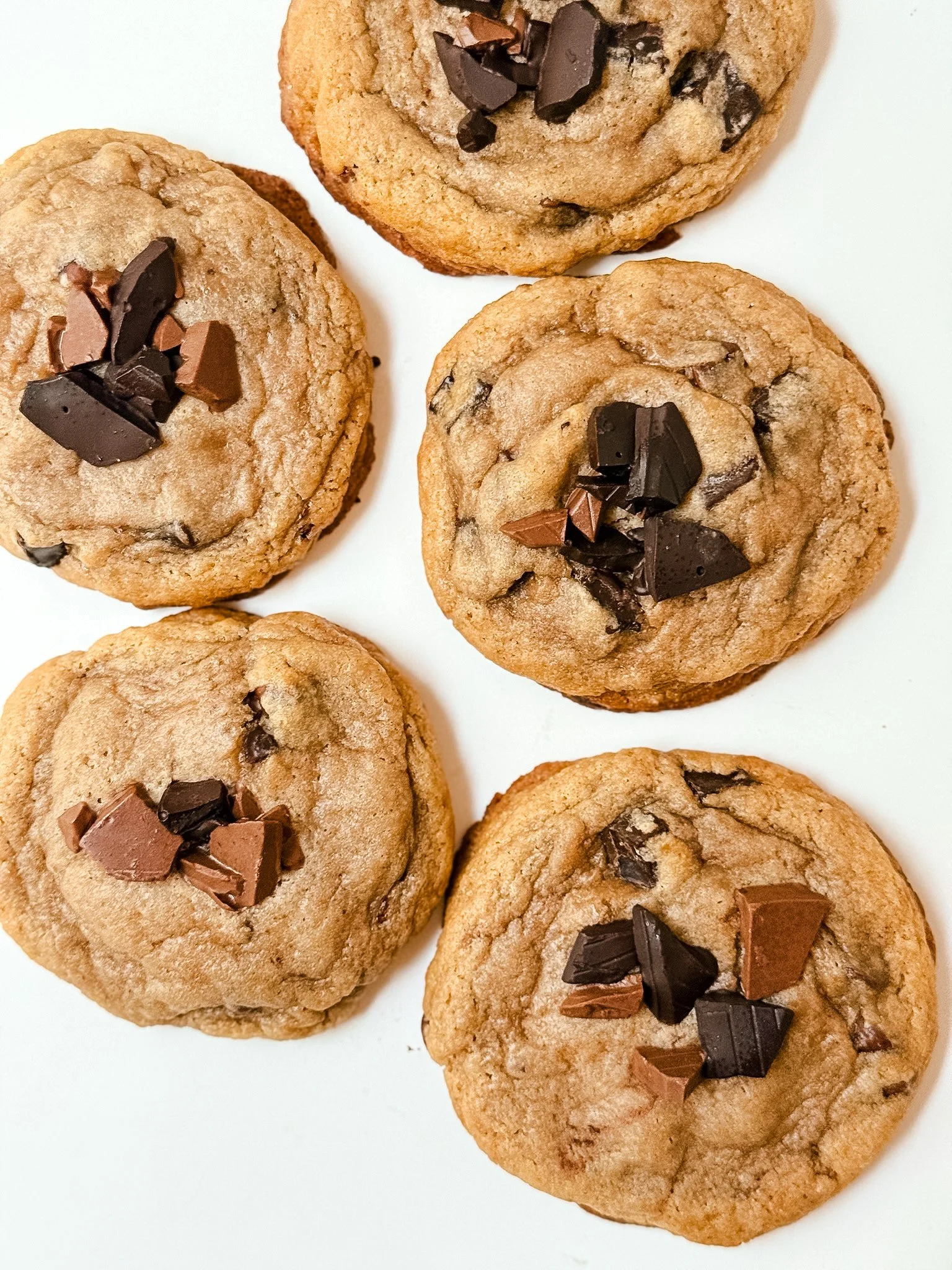 Several chocolate chip cookies with pieces of chocolate on top, arranged on a white surface.