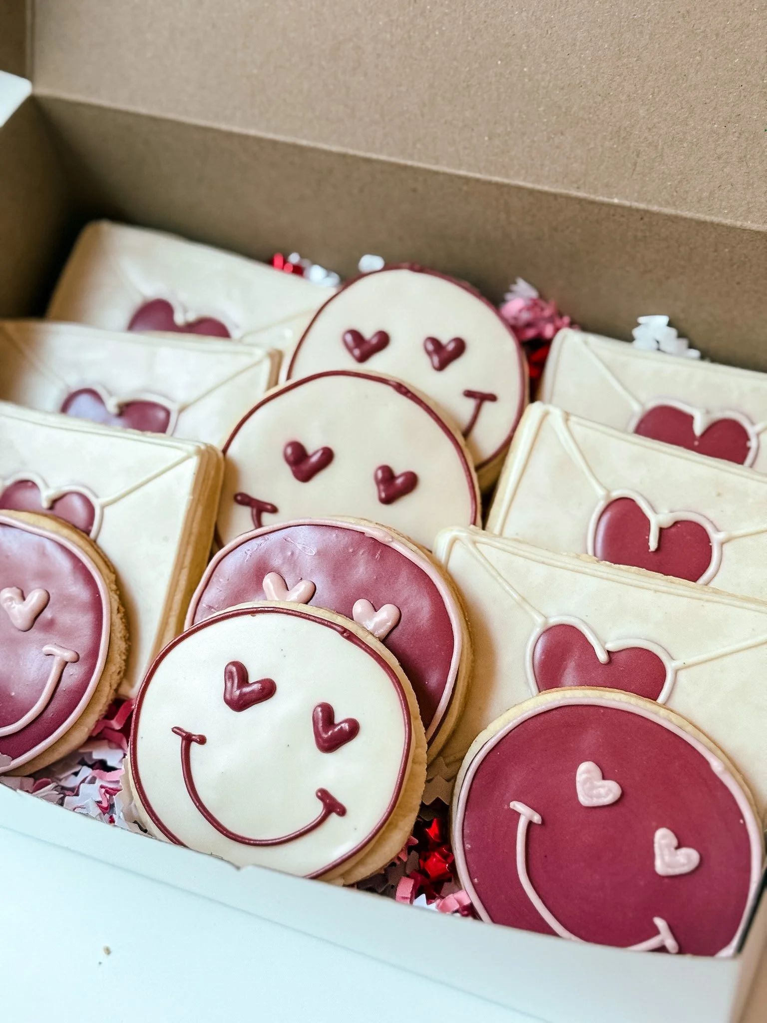 Box of decorated cookies with pink, white, and maroon frosting, some shaped like hearts and smiley faces, inside a white box lined with red and white shredded paper.