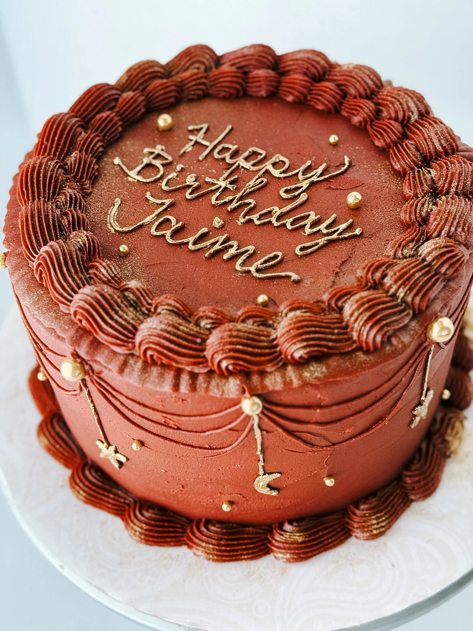 A round chocolate birthday cake with decorative borders made of piped chocolate frosting, gold edible beads, and the message 'Happy Birthday Jaimie' written on top in gold icing.