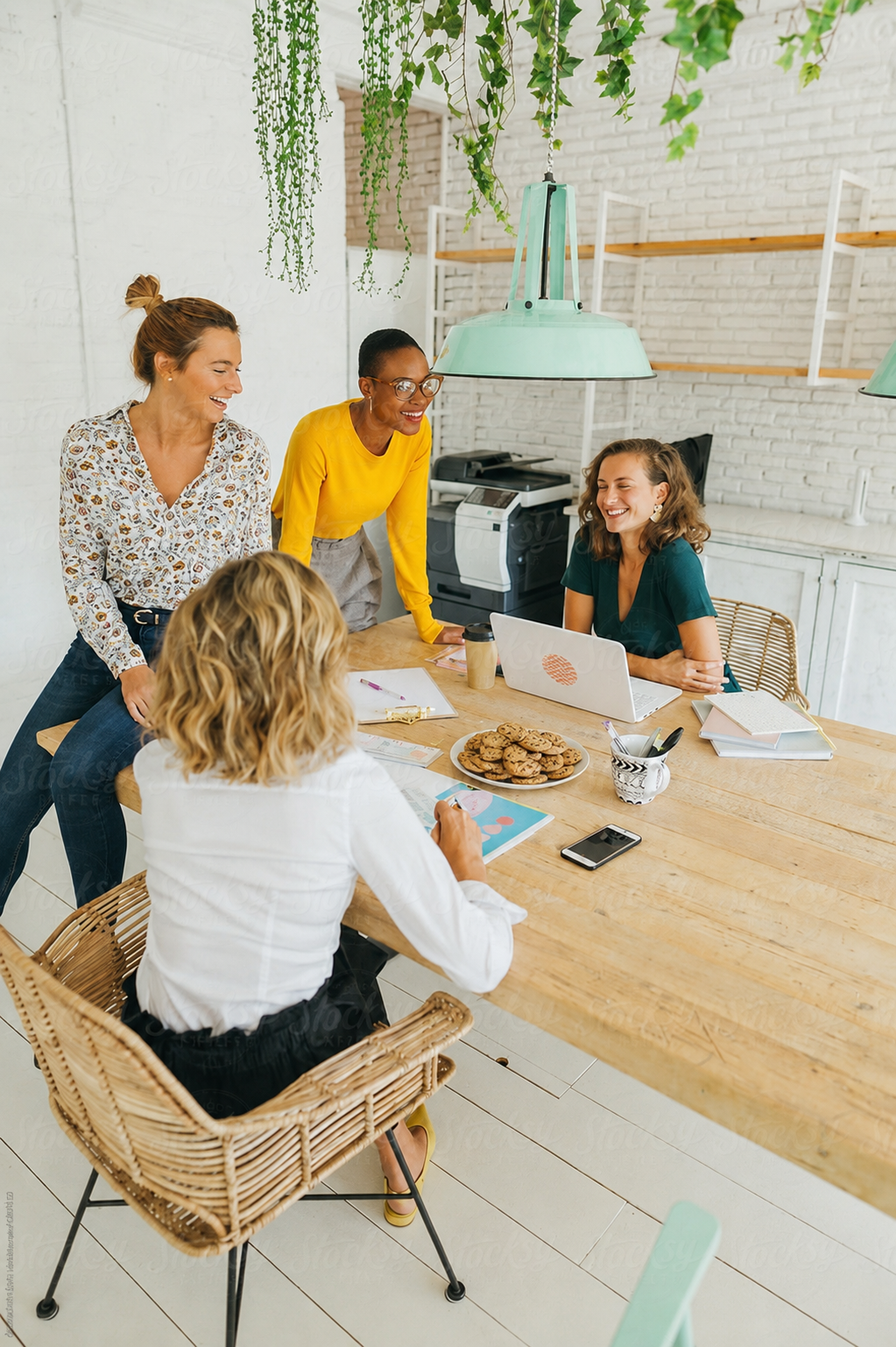 Four women gathered around a wooden table in a bright, modern office, laughing and talking. The table has a plate of cookies, notebooks, a smartphone, pens, and a laptop. Green plants hang from the ceiling.