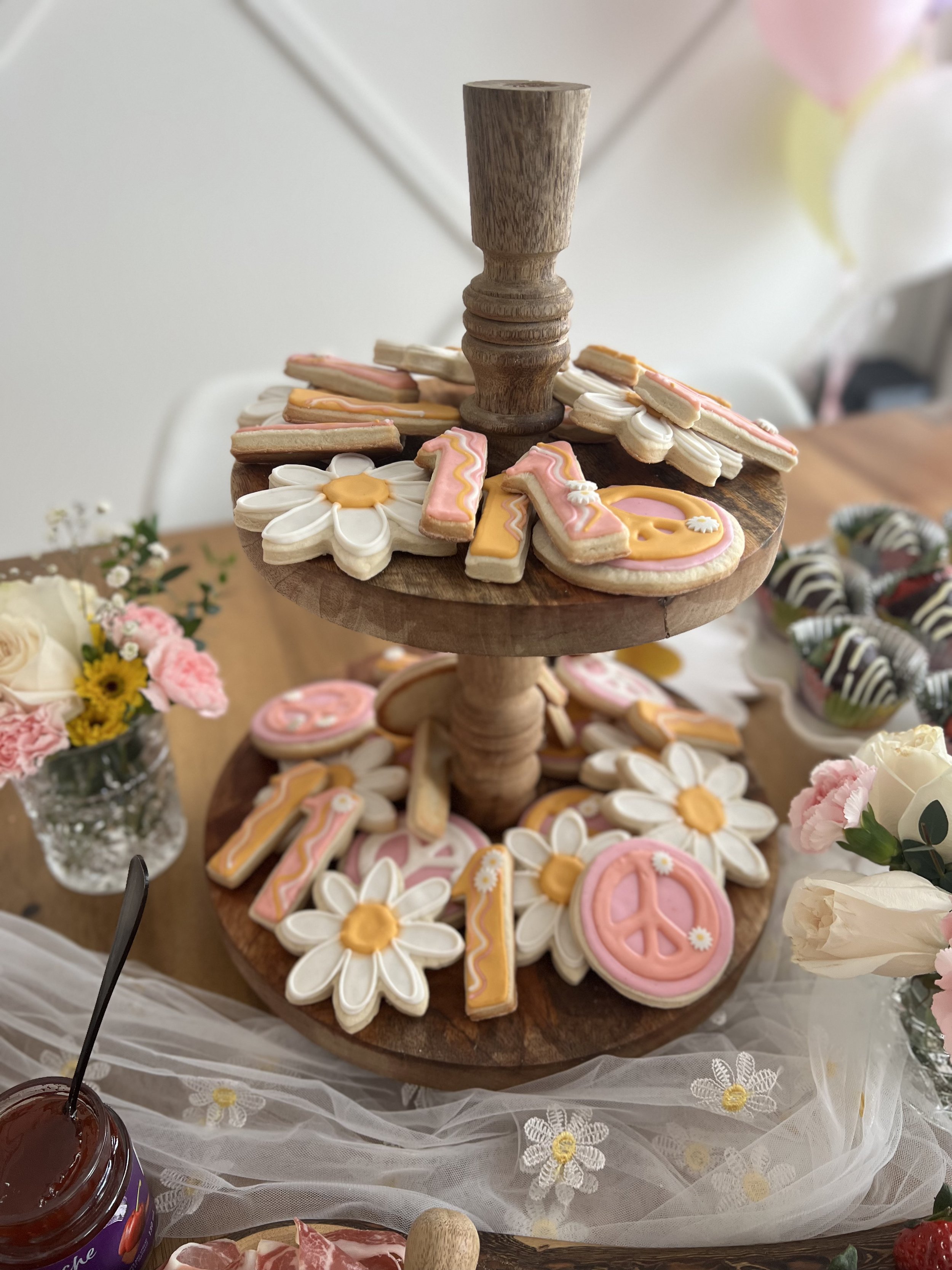 A wooden stand holding decorated cookies shaped like flowers, peace signs, and numbers, surrounded by flowers and a jar of jam.