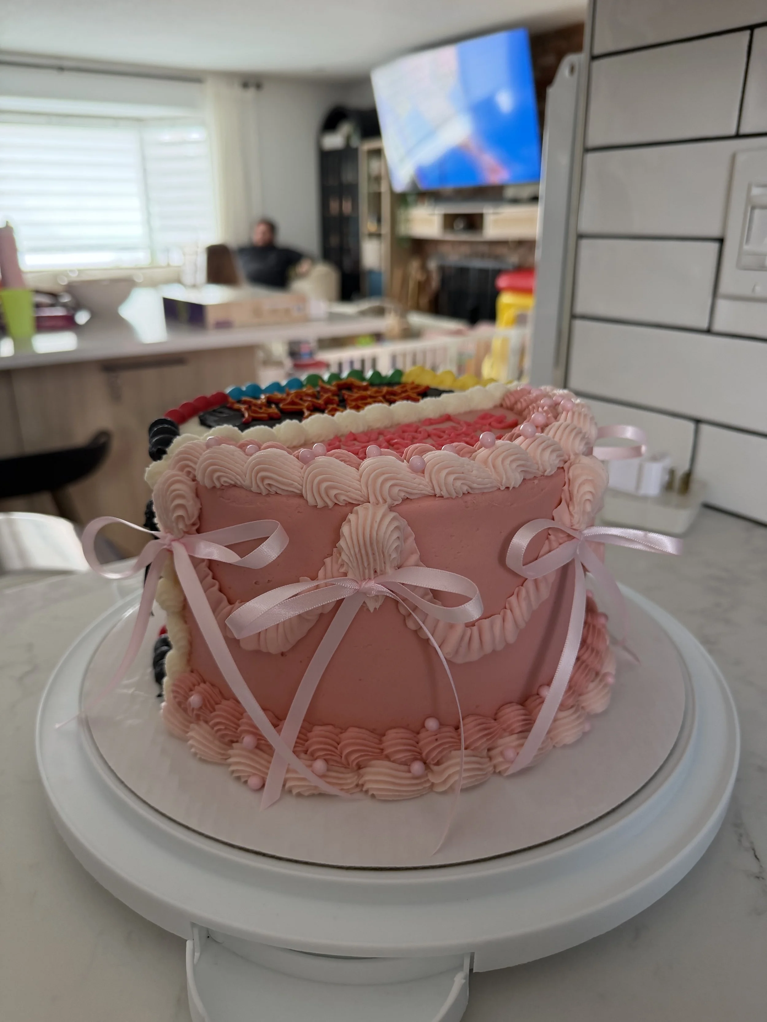 A pink birthday cake decorated with pink frosting, ribbons, and candy pearls, placed on a white cake stand.