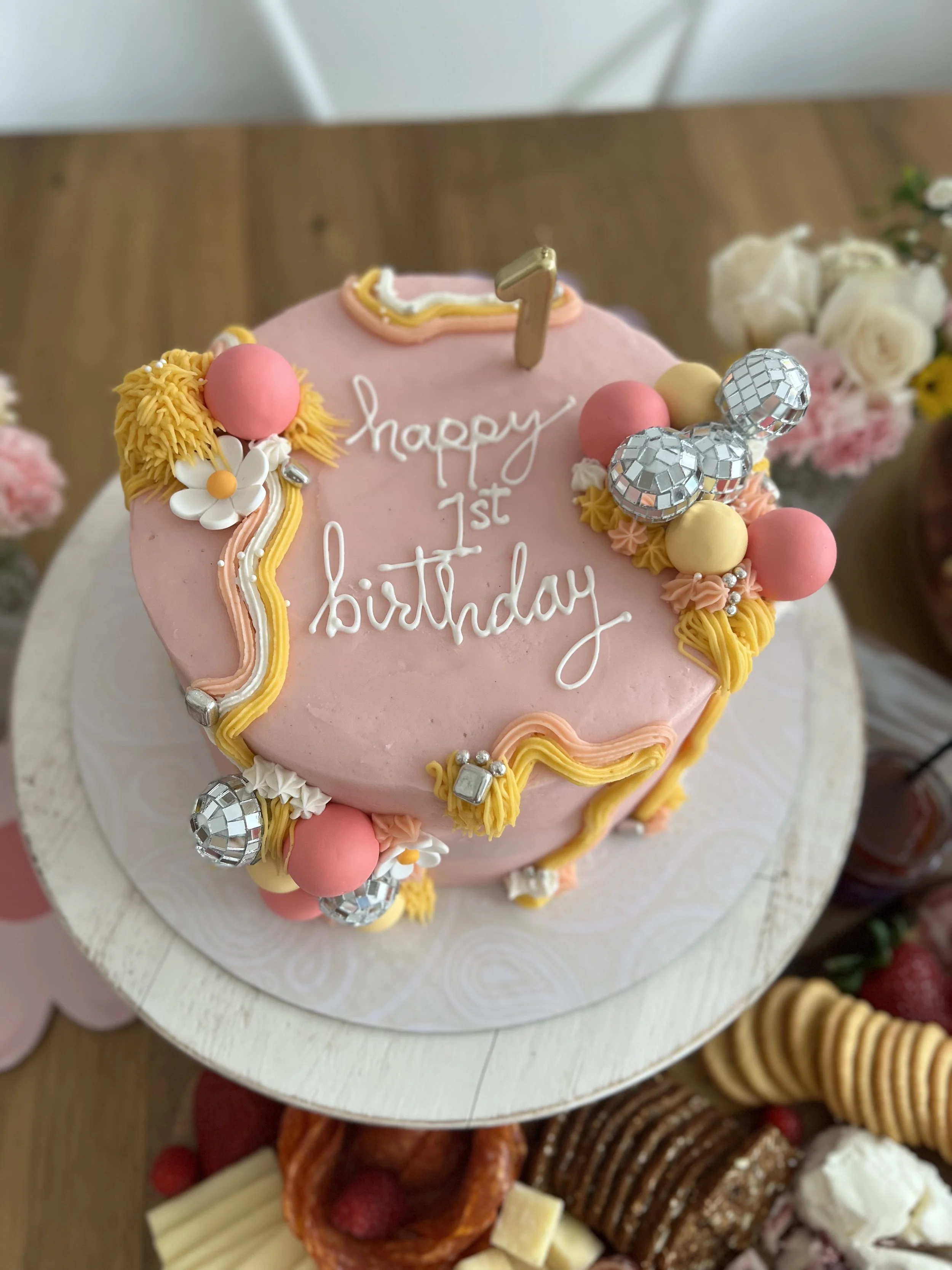 Pink birthday cake decorated with flowers, pink and yellow balls, and silver disco ball decorations. A gold number '1' topper and white cursive writing that says 'happy 1st birthday' are on the cake. The cake is on a white cake stand.