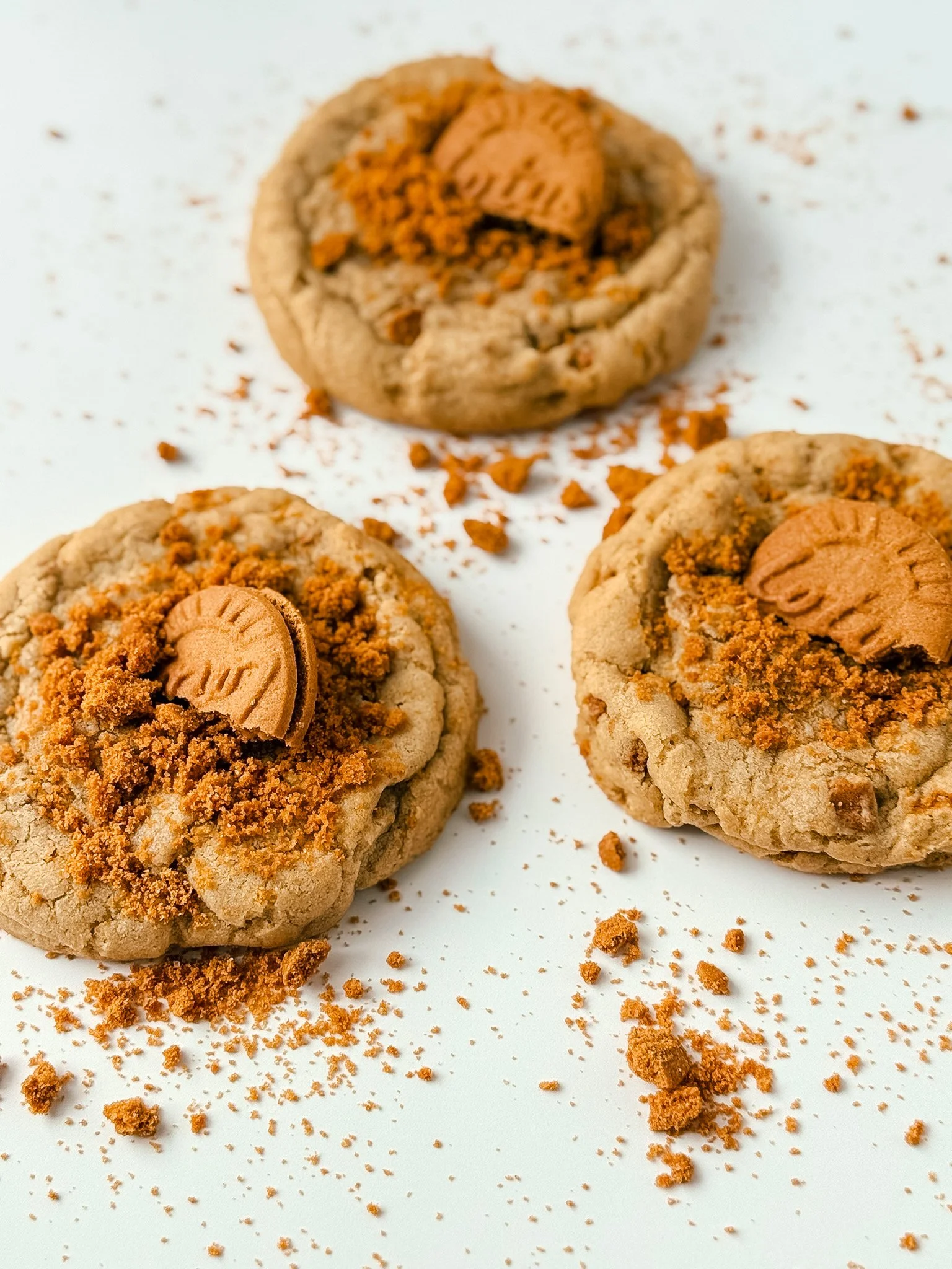 Close-up of three cookies topped with a shortbread cookie and crumbled cookie pieces, with crumbs scattered on a white surface.