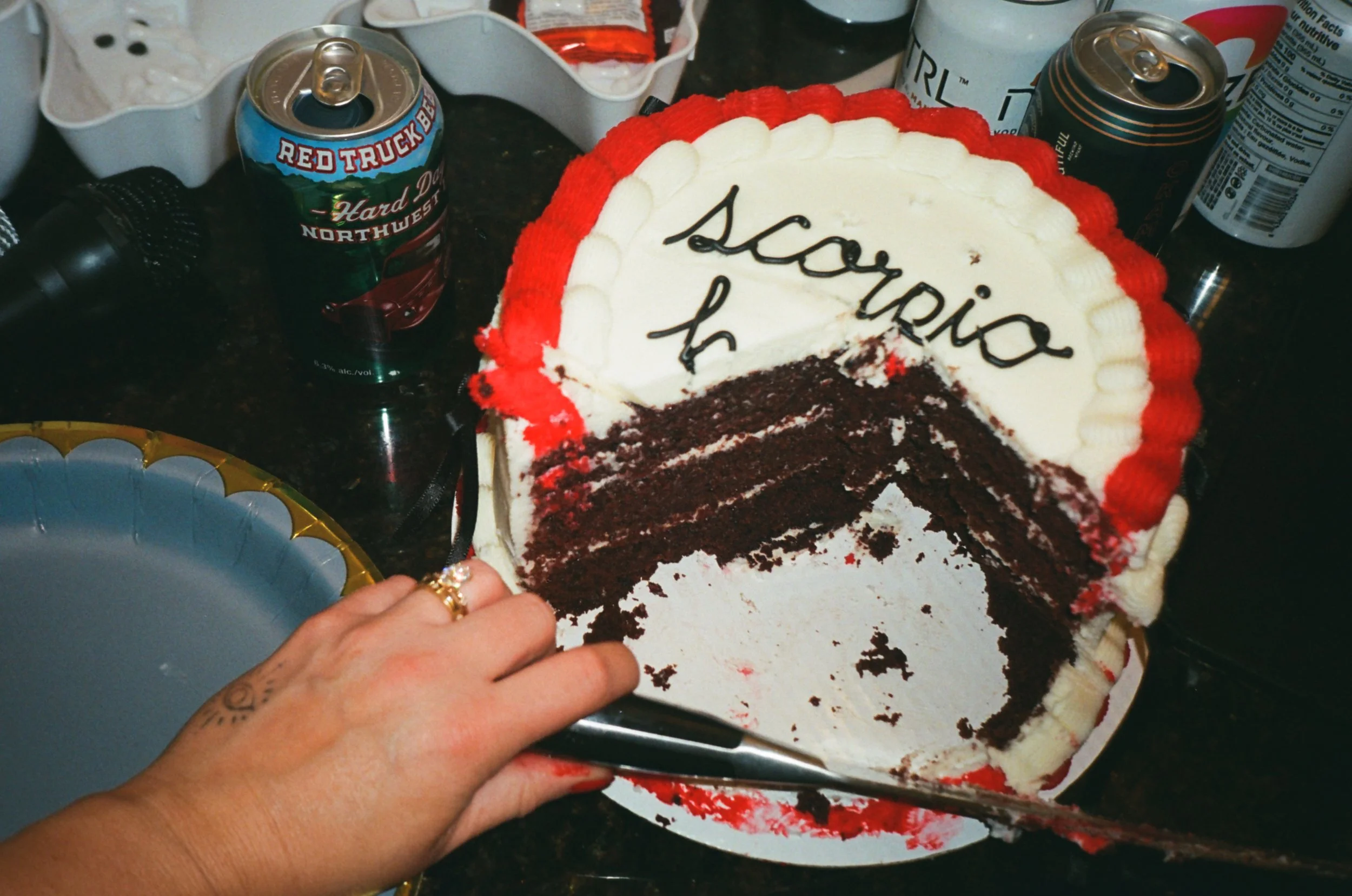 A birthday cake with red and white frosting, with the message 'Scooter H' written on top, surrounded by empty soda cans and party decorations.