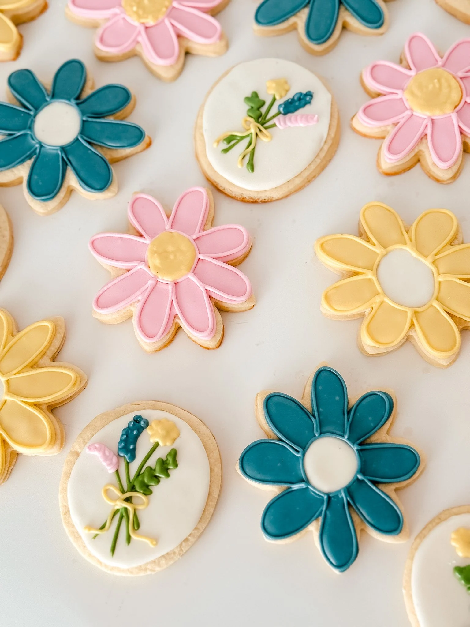 Colorful flower-shaped cookies decorated with pink, blue, yellow, and white icing, some with floral or bow designs, on a white surface.