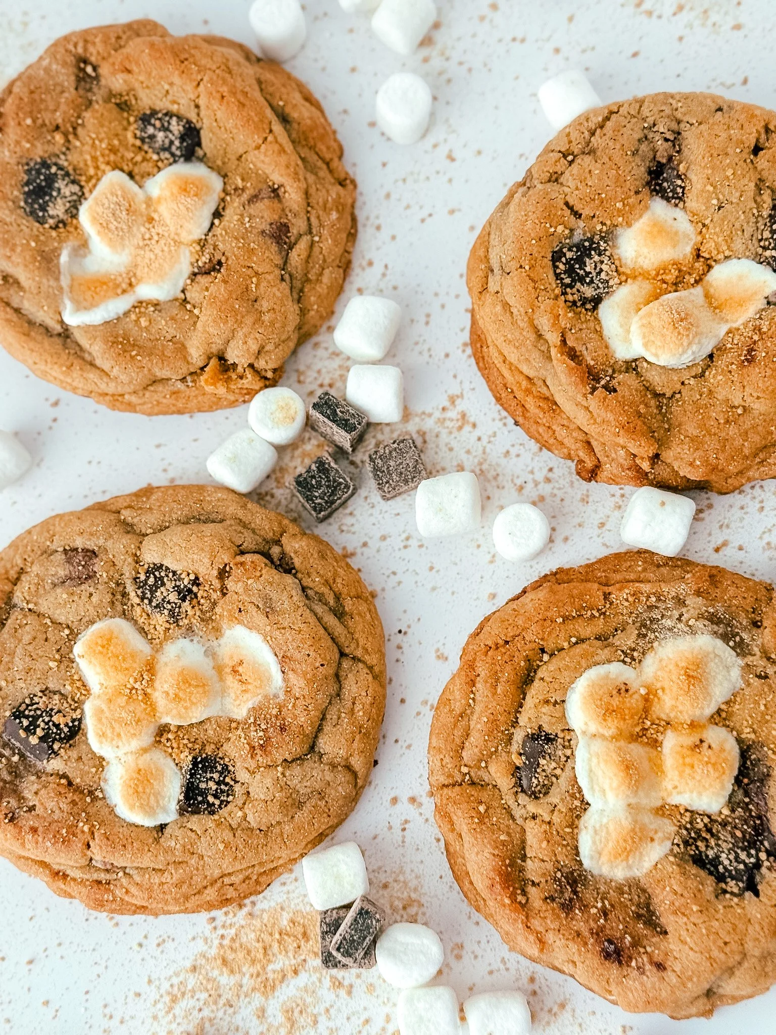 Close-up of three chocolate chip cookies decorated with toasted marshmallows and sprinkled with graham cracker crumbs, marshmallows, chocolate pieces, and graham crackers scattered around on a white surface.