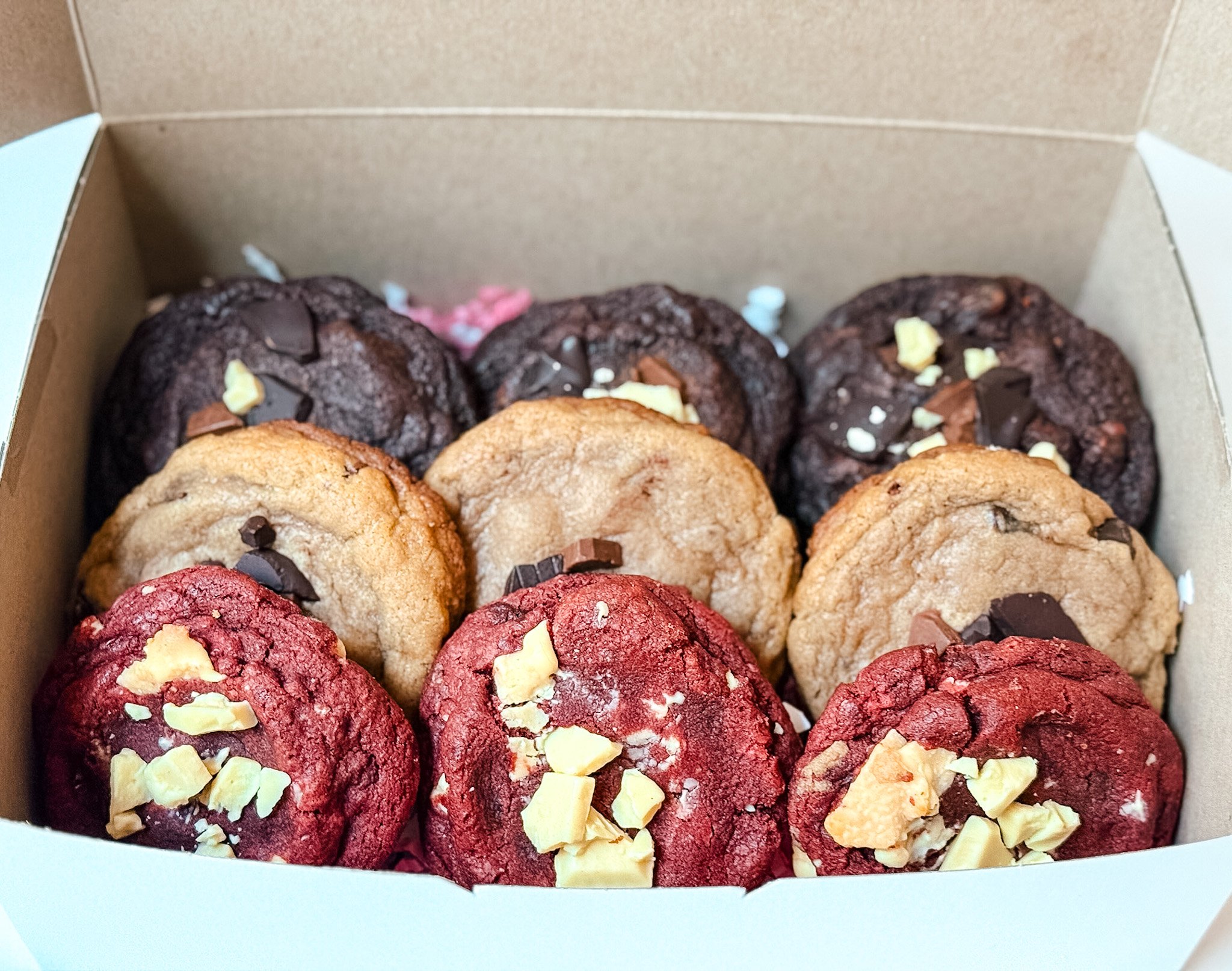 A close-up view of a box with assorted cookies, including chocolate and berry-flavored cookies topped with white chocolate chunks.