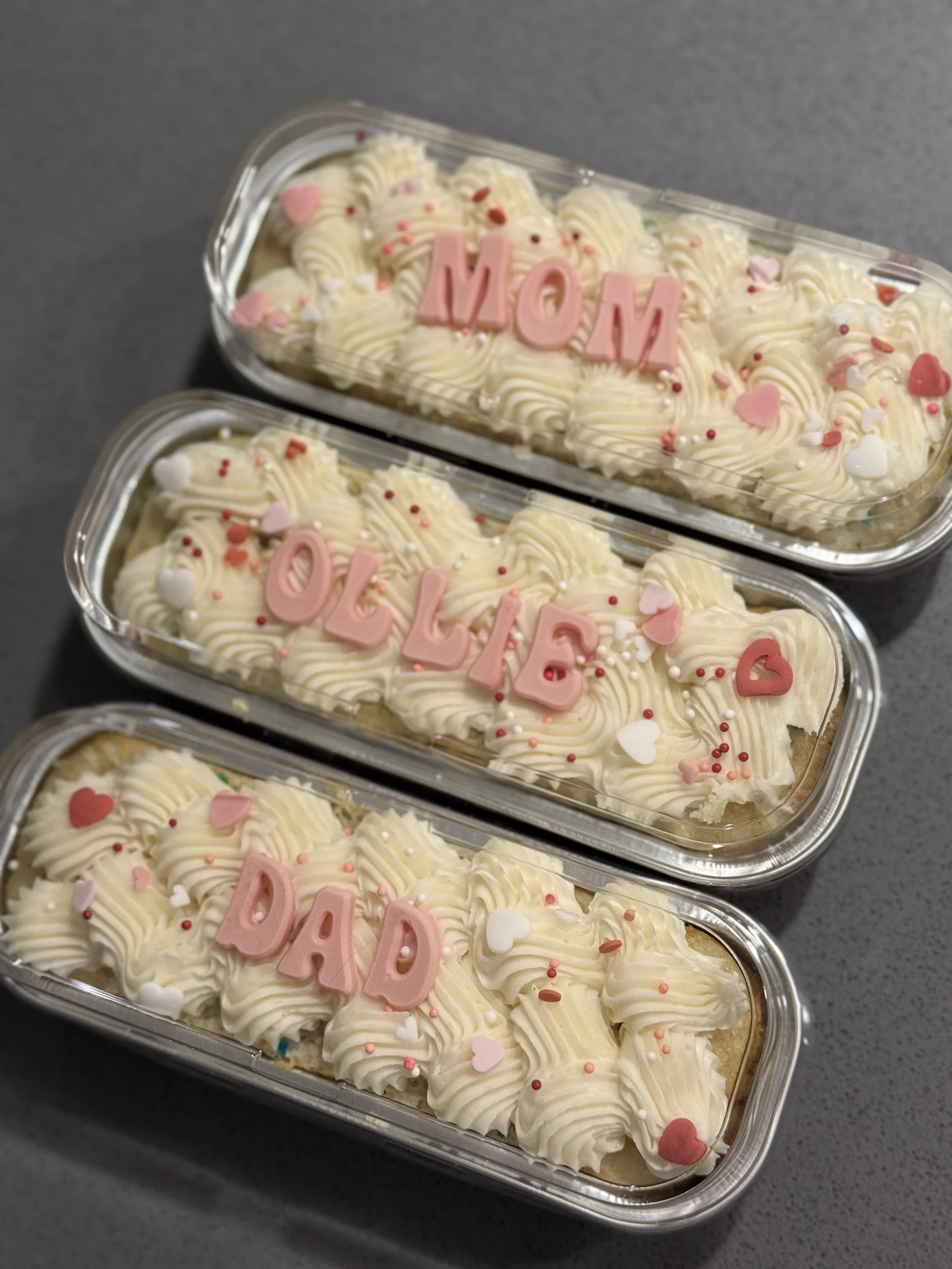 Two dessert containers with white rose-shaped frosting, pink and white icing hearts, and multicolored sprinkles. Pink letters spell 'MOM' on each container.