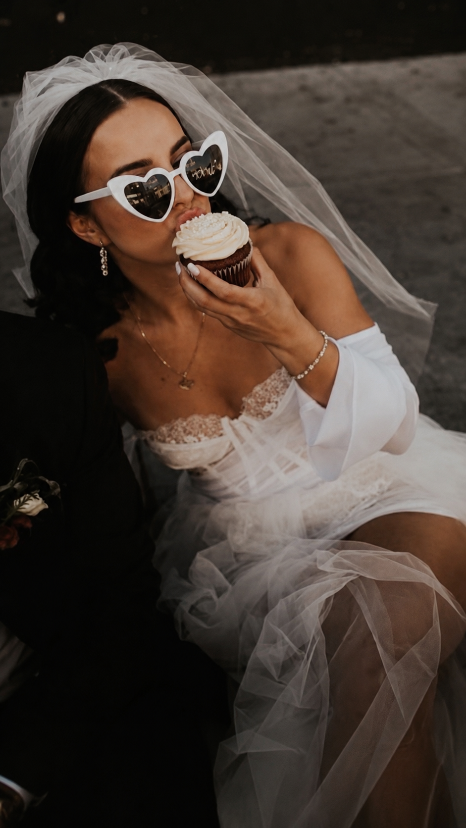 Woman in a wedding dress with heart-shaped sunglasses eating a cupcake.