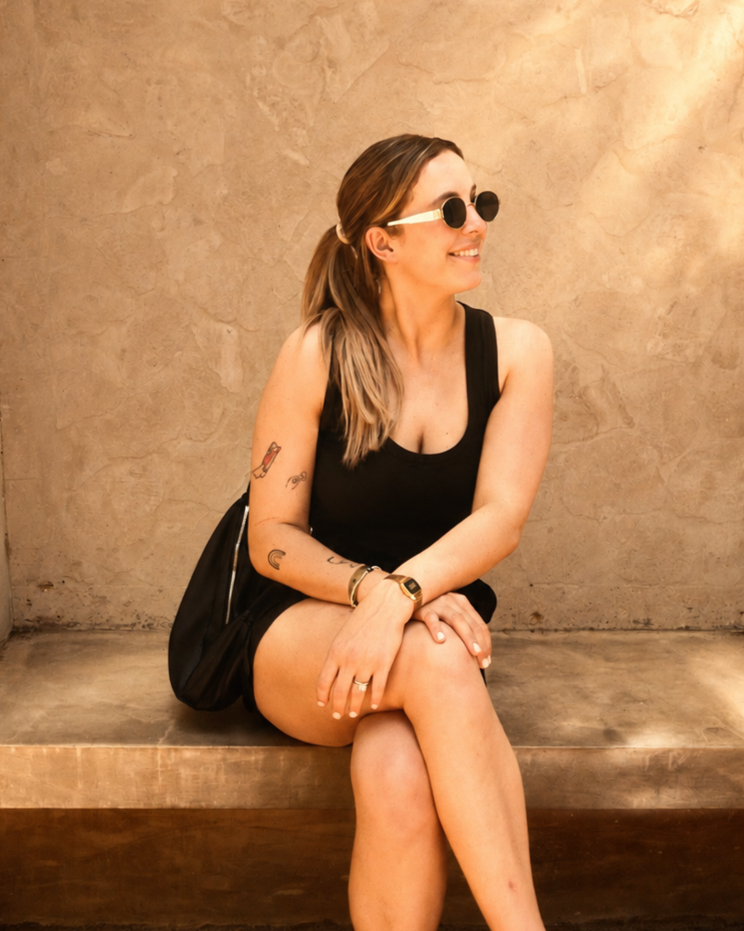 A woman with long hair sitting on a wooden bench against a beige wall, wearing sunglasses, a sleeveless black top, and shorts, smiling and looking to her left.