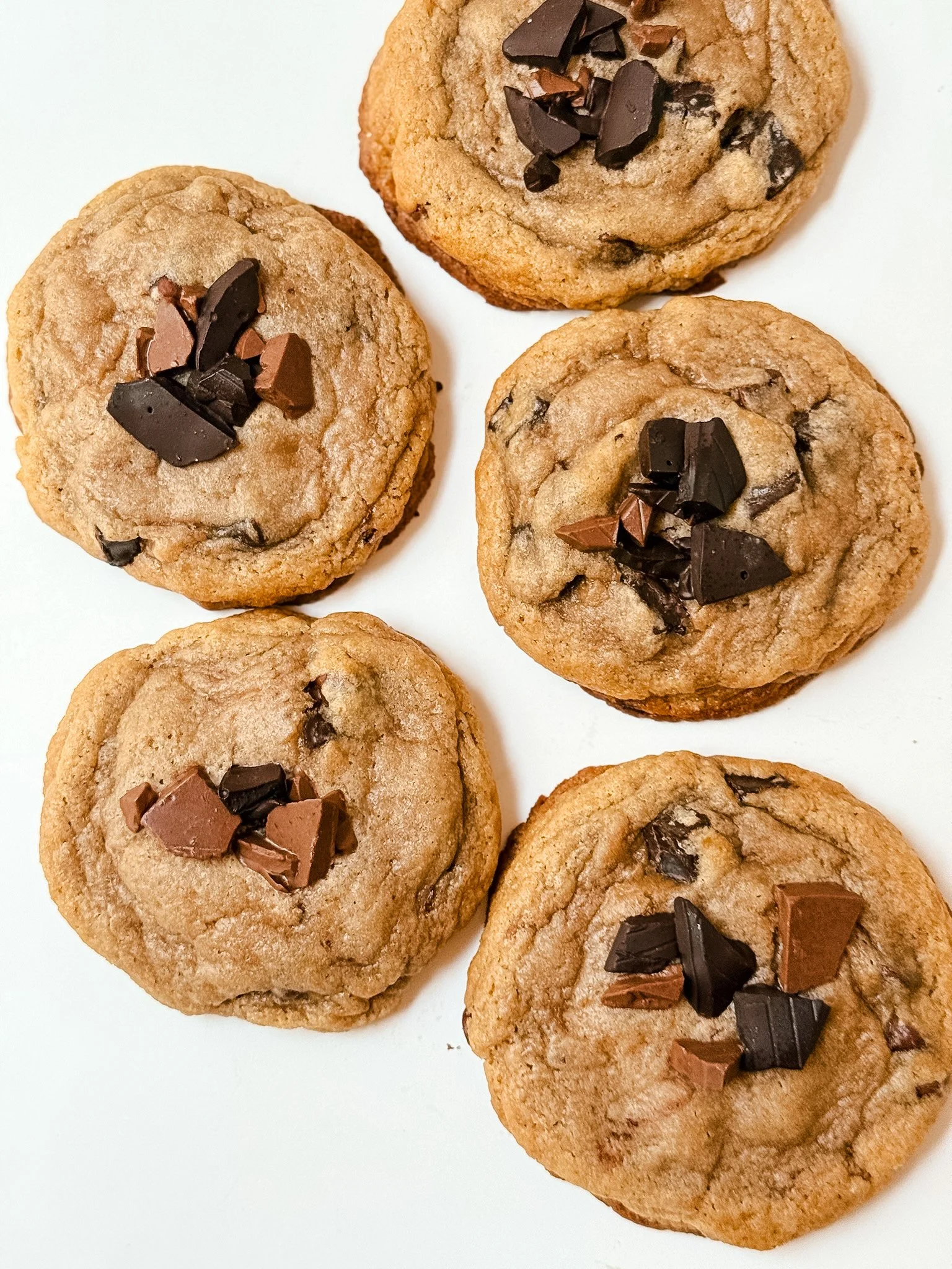 Close-up of six chocolate chip cookies with chunks of dark and milk chocolate, placed on a white surface.