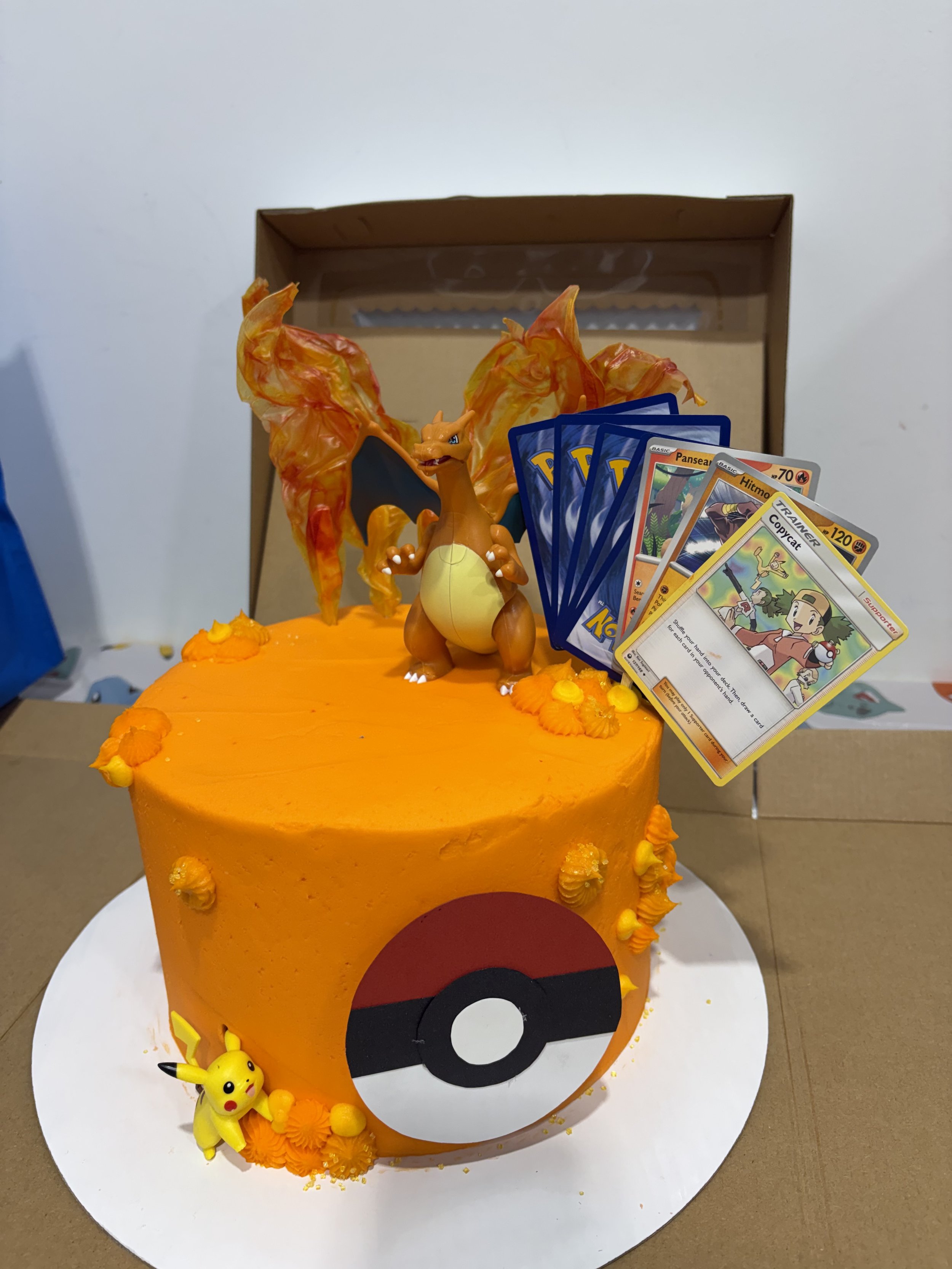 A decorative cake with Pokémon theme featuring a Charizard figure, Pikachu, and Pokémon cards on top, decorated with orange and yellow frosting.