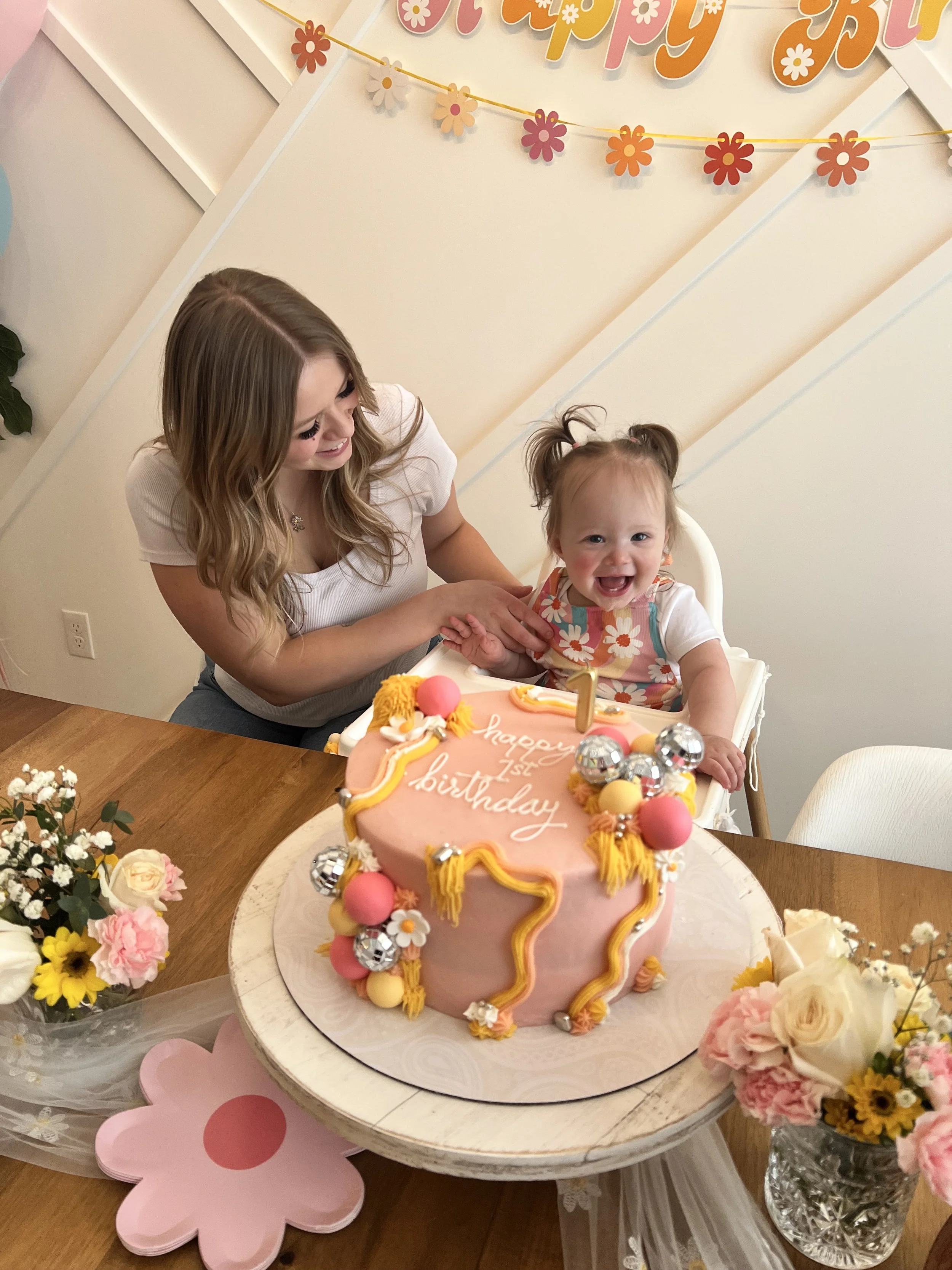 A child and woman smiling at a birthday celebration with a pink and yellow decorated cake with the words 'happy 1st birthday' written on it. Flowers and decorations surround the scene.